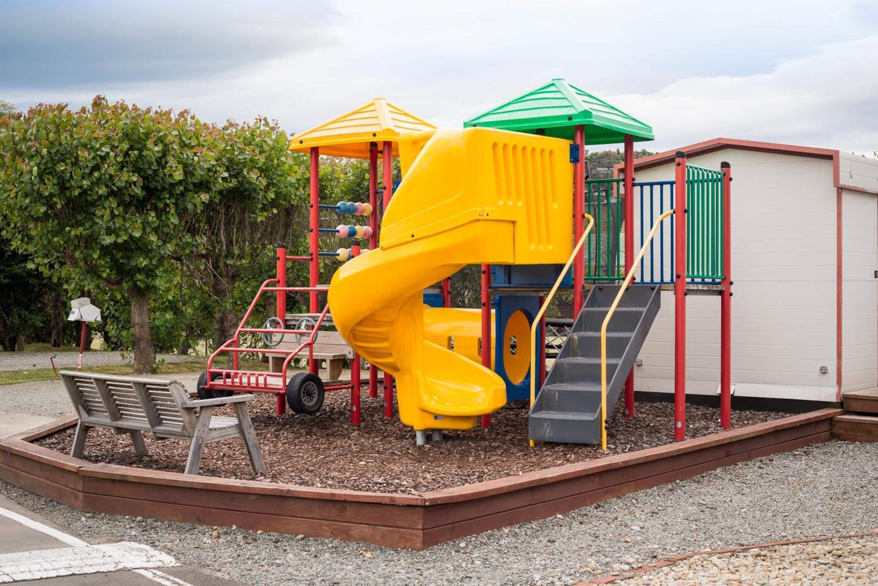 Une aire de jeux avec un toboggan jaune, une cabane de jeux dotée d’un toit vert et un escalier gris.