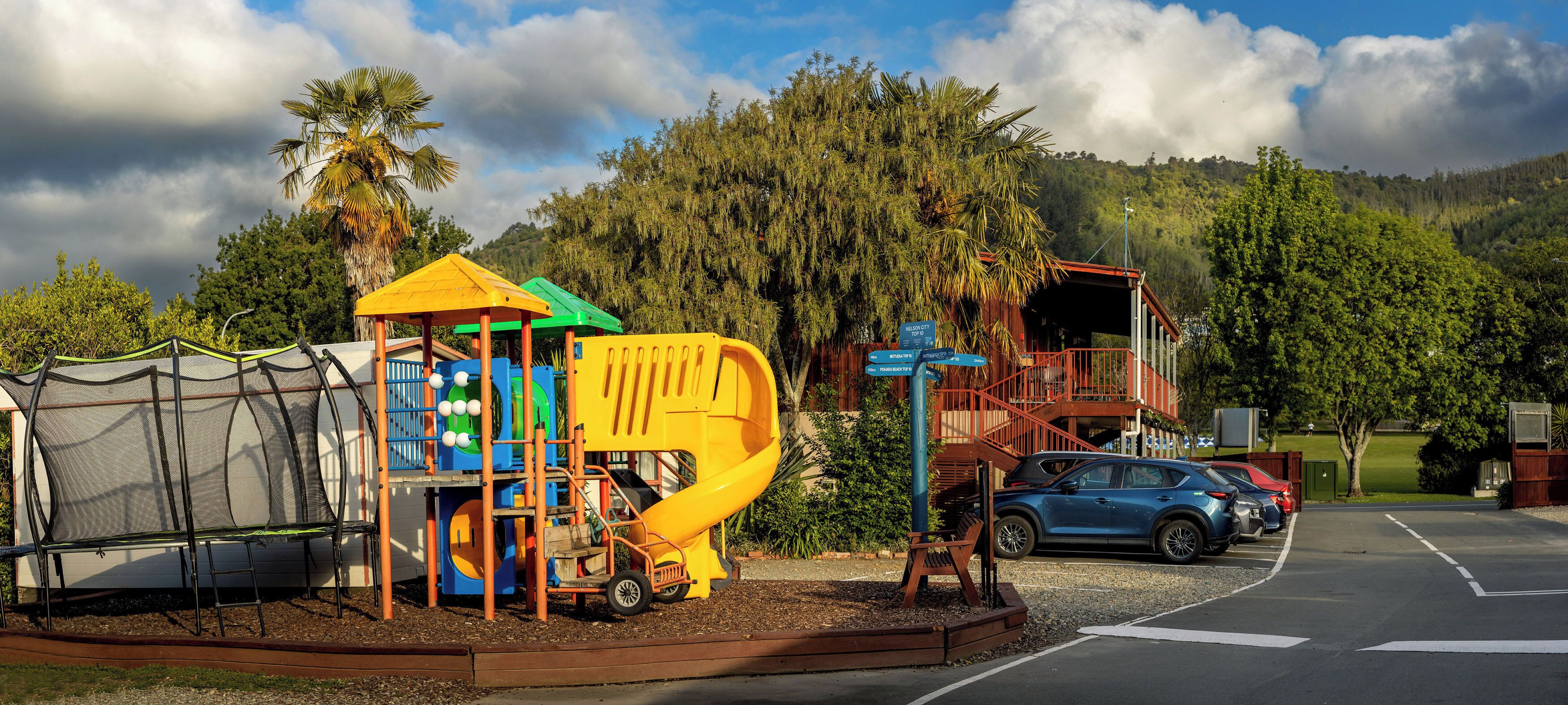 A playground with a slide and climbing structures, surrounded by a parking area and a building with a red roof.