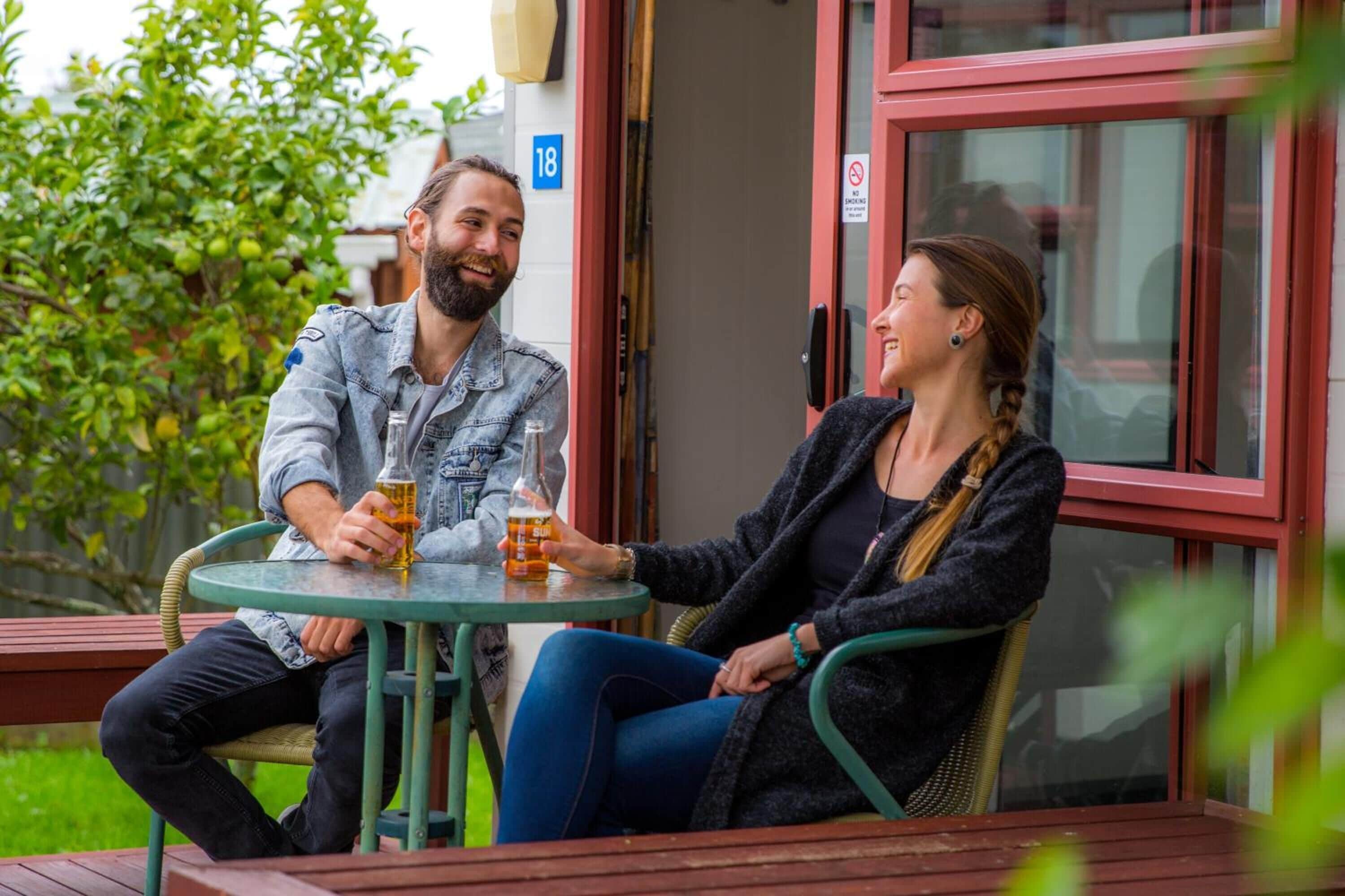 Two people sitting at an outdoor table with drinks, a building number 18, and a no smoking sign.