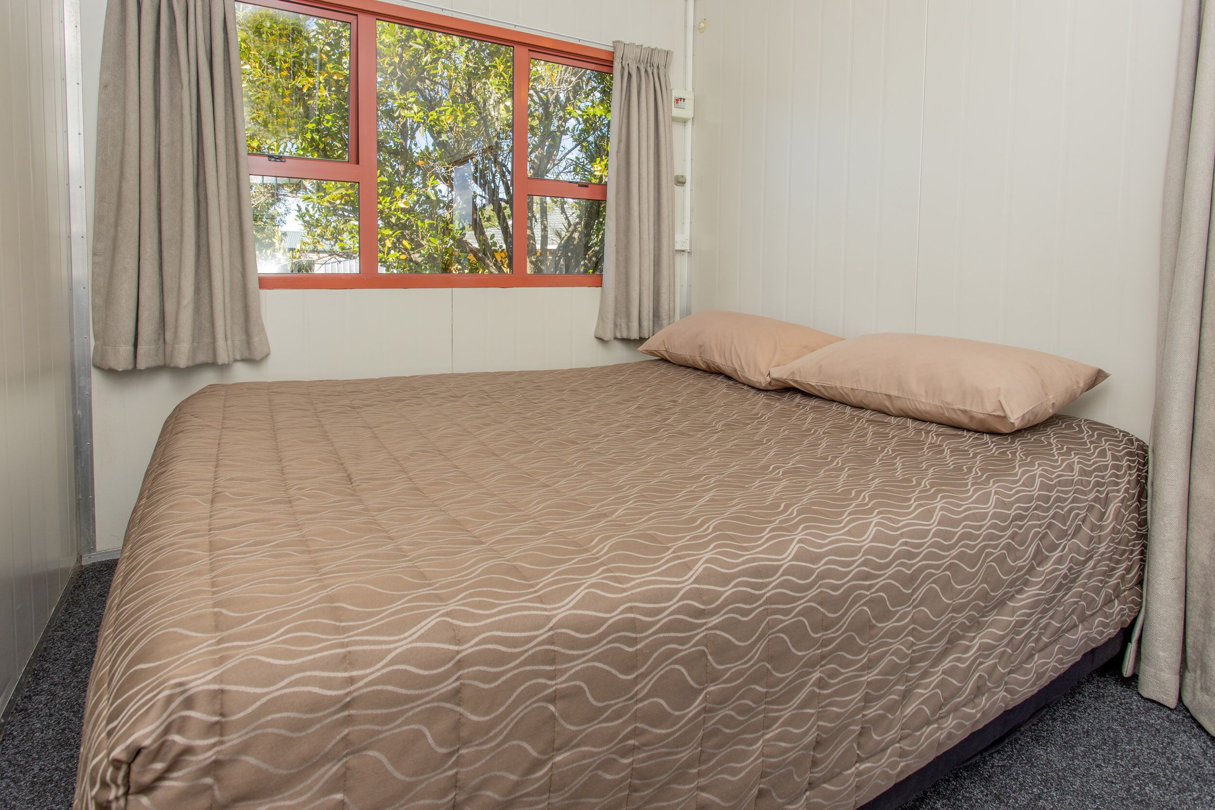 Una cama bien tendida con una colcha estampada, una ventana con marco rojo y cortinas.