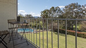 A balcony with a laptop, a table, and chairs, overlooking a swimming pool and a well-maintained lawn with trees and bushes. - Harriet House Tumut (Tumut)