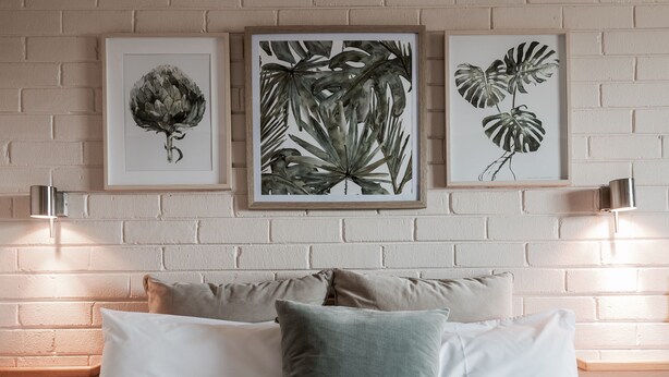 A bed with white bedding and multiple pillows, flanked by two wall-mounted light fixtures and three framed botanical prints on a brick wall. - Harriet House Tumut (Tumut)
