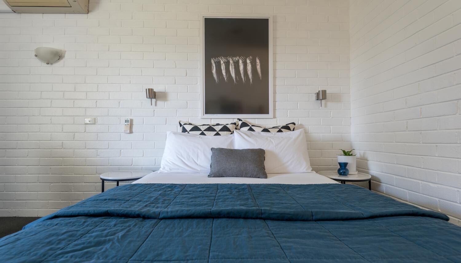 Un dormitorio con cama, paredes de ladrillo blanco, almohada negra y blanca, mesita de noche con una planta, y un aplique de pared.