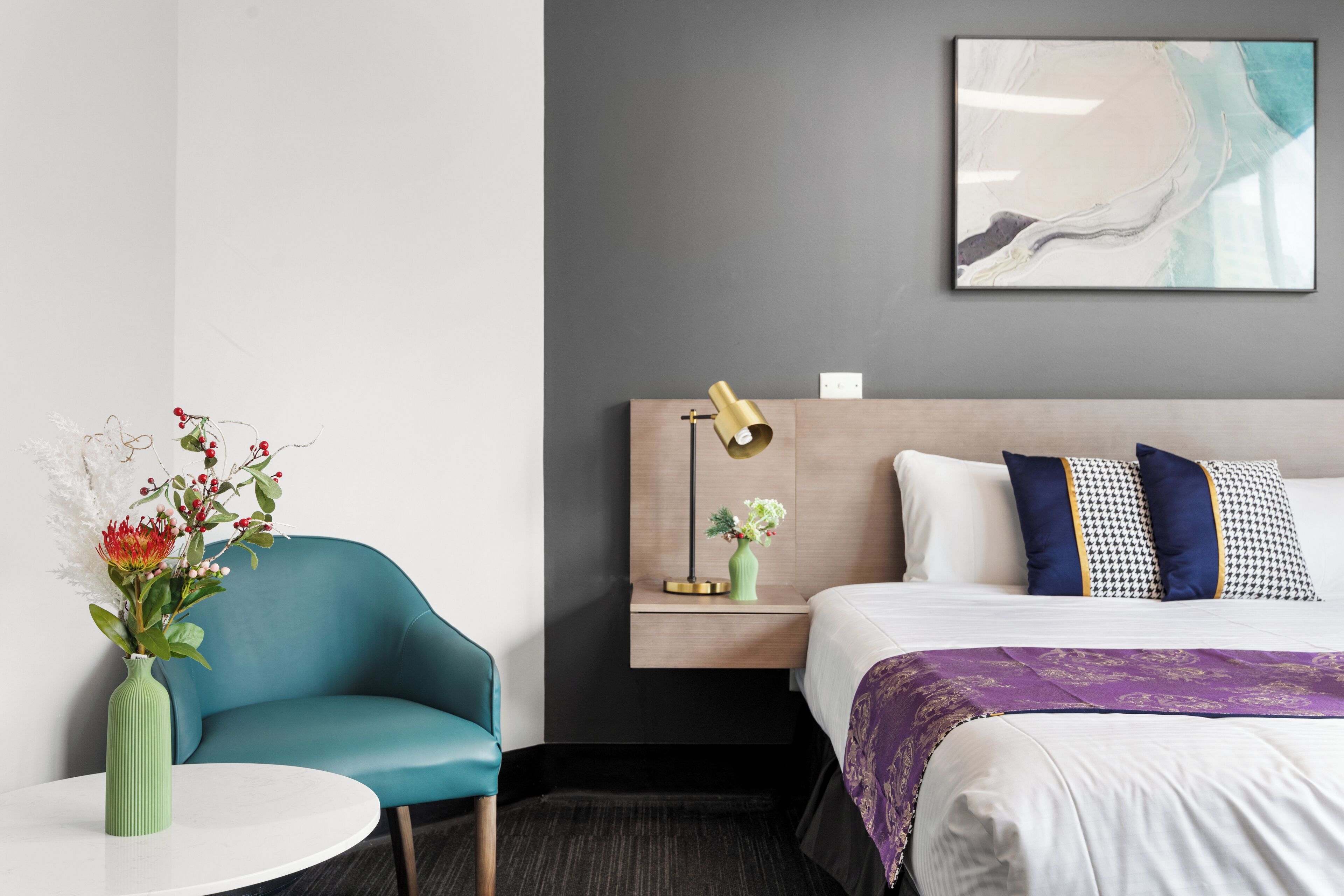 Ein Hotelzimmer mit einem petrolfarbenen Sessel, einem runden weißen Tisch mit einer grünen Vase, einem Bett mit weißer und violetter Bettwäsche, einem Kopfteil mit einer Lampe sowie einem an der Wand hängenden abstrakten Gemälde.