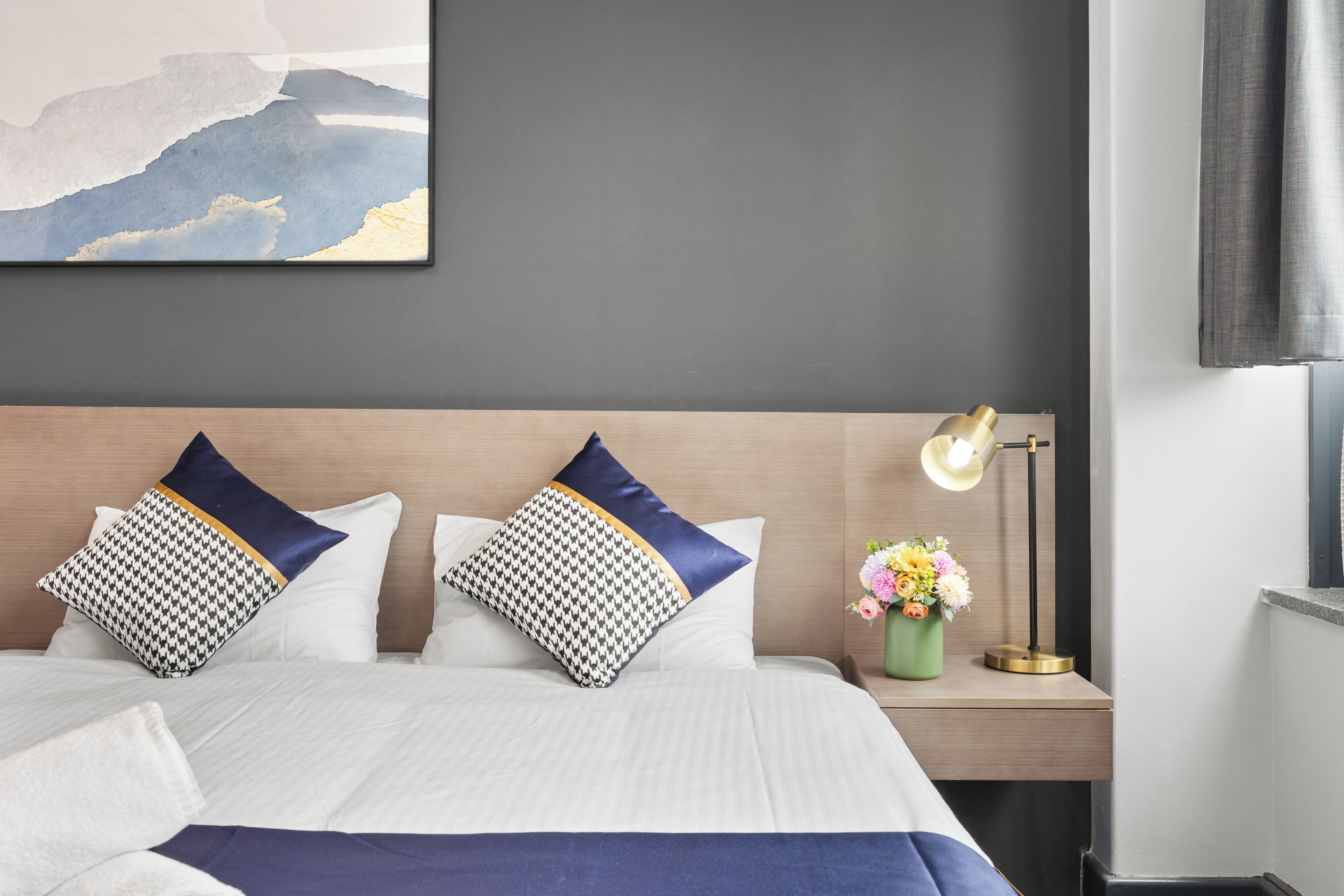 Una cama bien tendida con ropa blanca y dos almohadas estampadas, una mesita de noche con una lámpara y un jarrón con flores, y un cuadro abstracto grande en la pared.