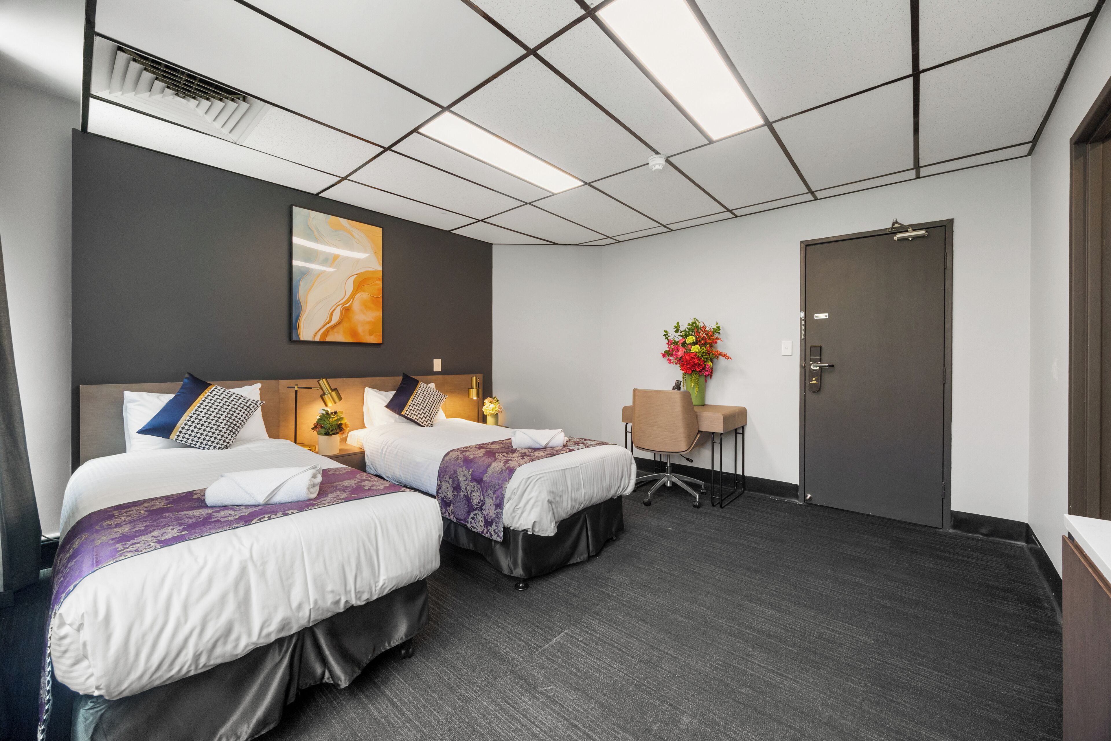 A hotel room with two beds, a desk, a chair, a door, and a painting on the wall.