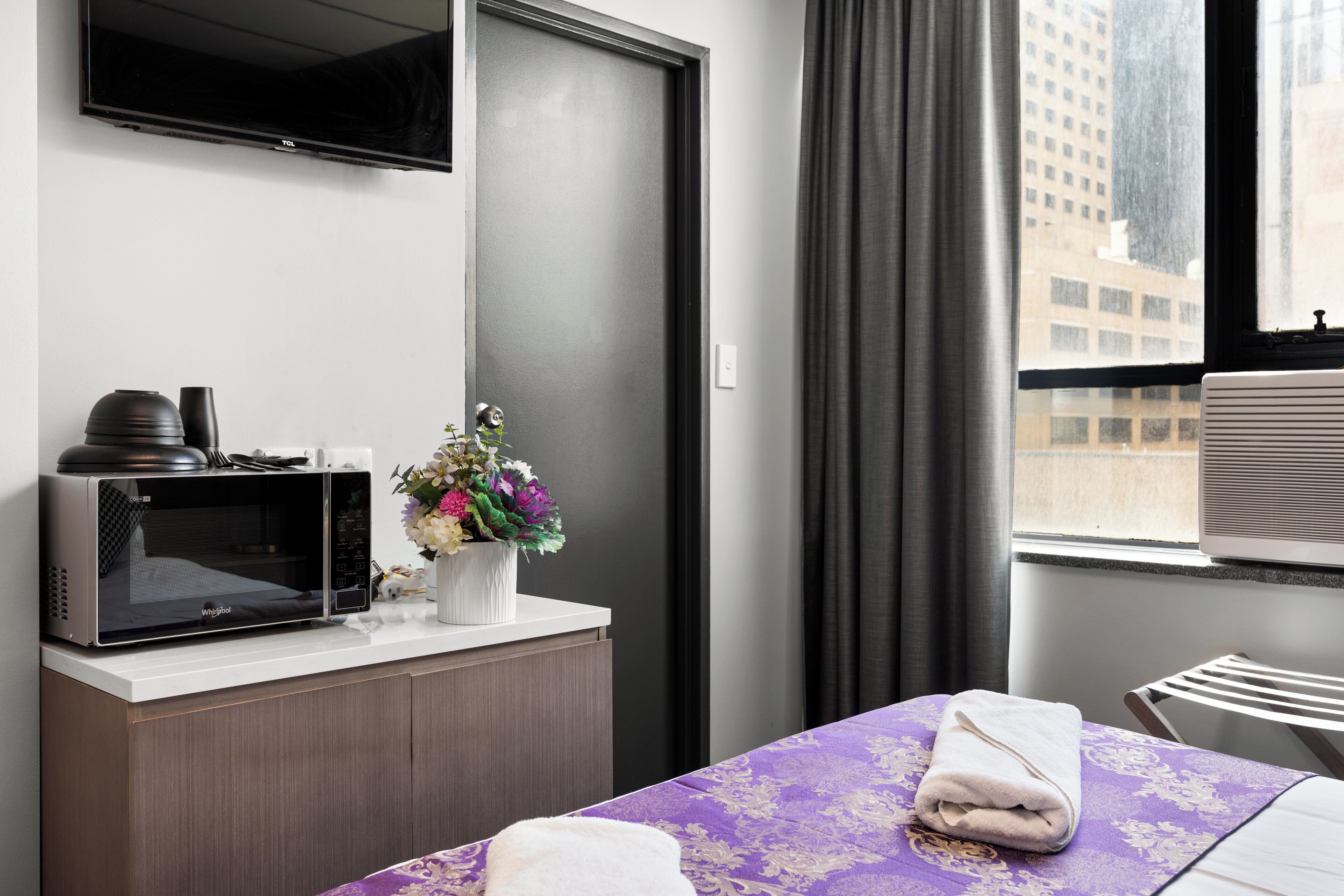 Ein Hotelzimmer mit einer Mikrowelle auf einer Arbeitsfläche, einer Vase mit Blumen, einem Bett mit lila Bettdecke und einem Fenster mit Blick auf Gebäude.
