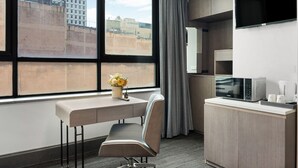 A hotel room with a desk, chair, kitchenette, and a large window with a view of buildings. - Eminence on Bank (Adelaide)