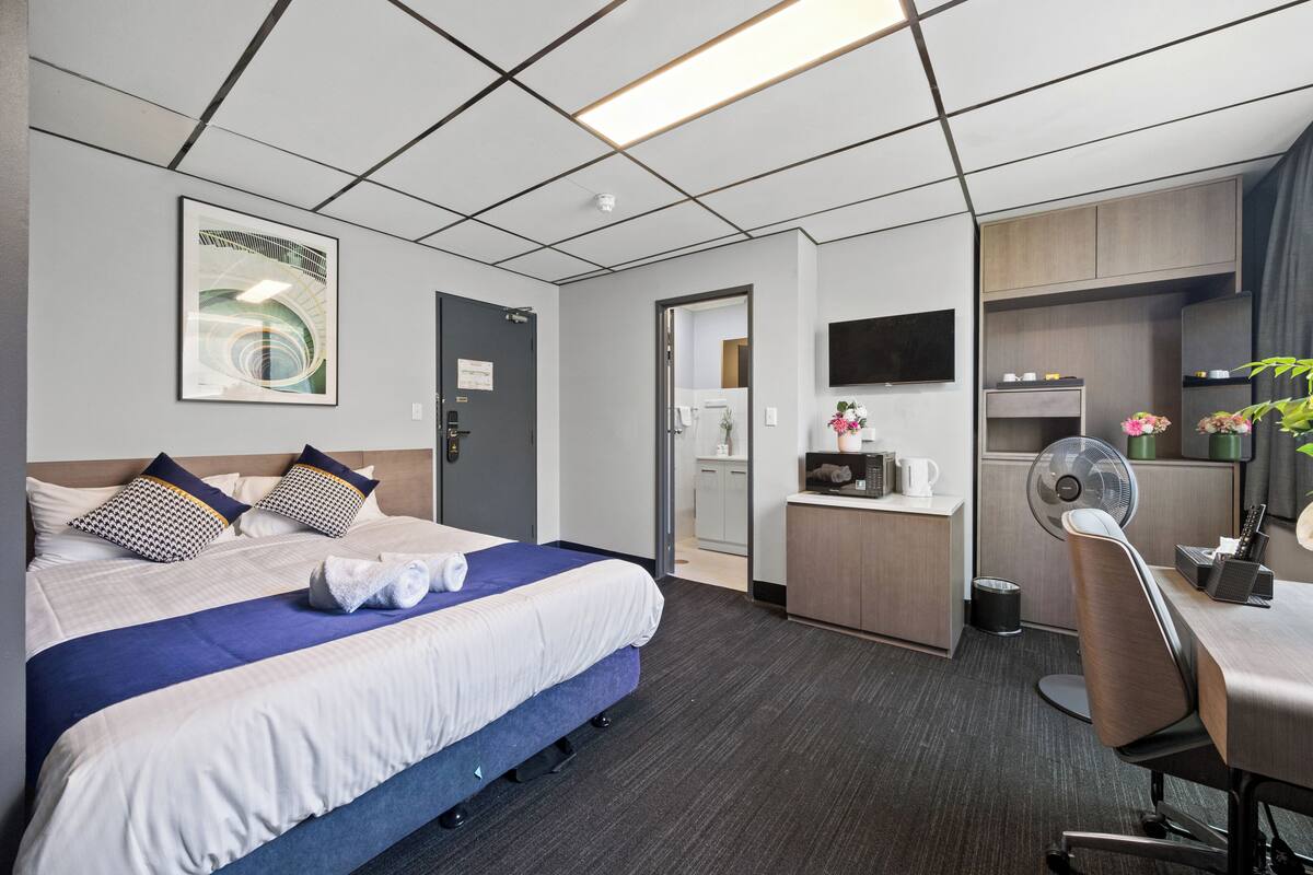 A hotel room with a bed, a desk, a chair, a TV, and a kitchenette.