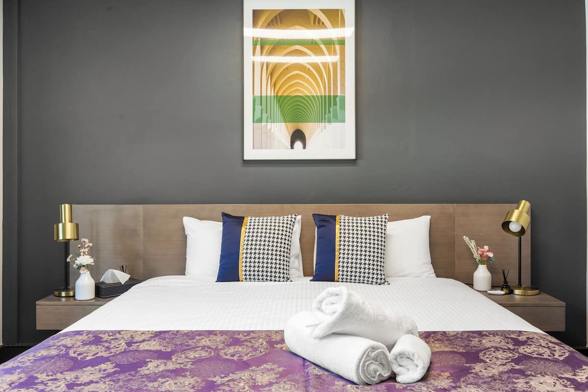 A neatly made bed with a purple and gold bedspread, two patterned pillows, a white vase with flowers, and a framed abstract artwork above the bed.