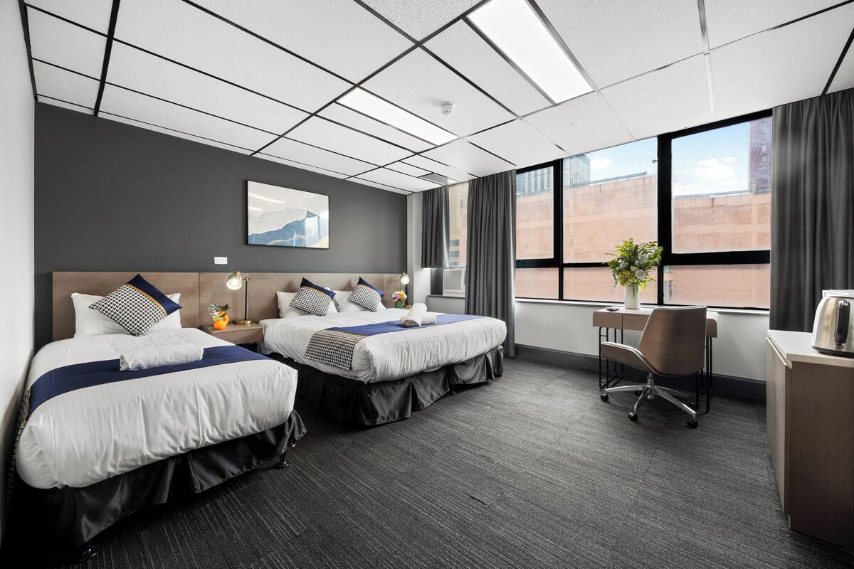 A hotel room with two beds, a desk, a chair, a window with curtains, and a wall-mounted artwork.