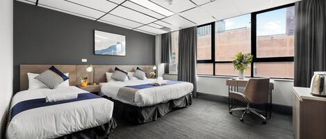 A hotel room with two beds, a desk, a chair, a window with curtains, and a wall-mounted artwork.
