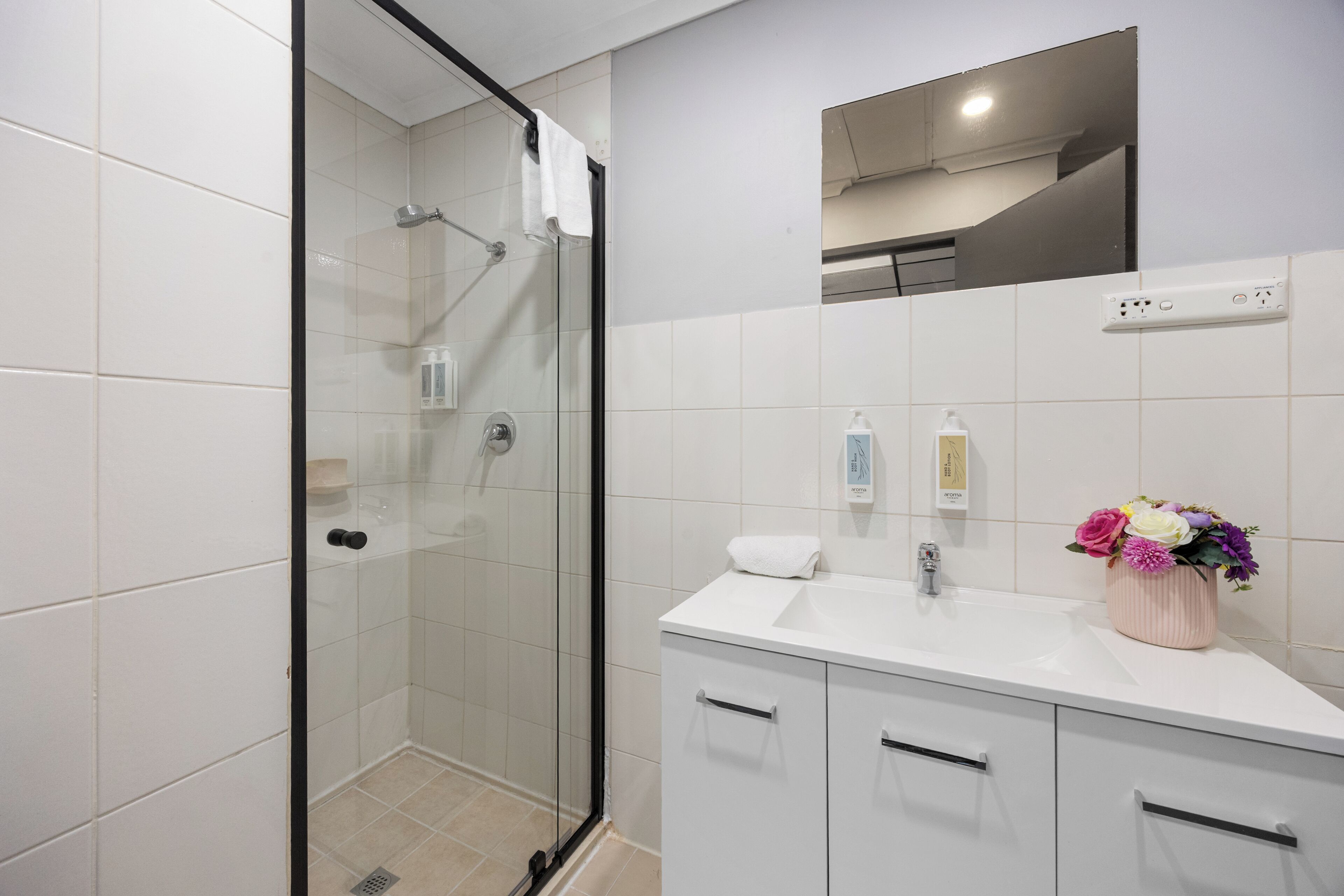 Une salle de bain moderne avec une douche à parois de verre, un lavabo blanc agrémenté d’un vase de fleurs et un miroir au-dessus du lavabo.
