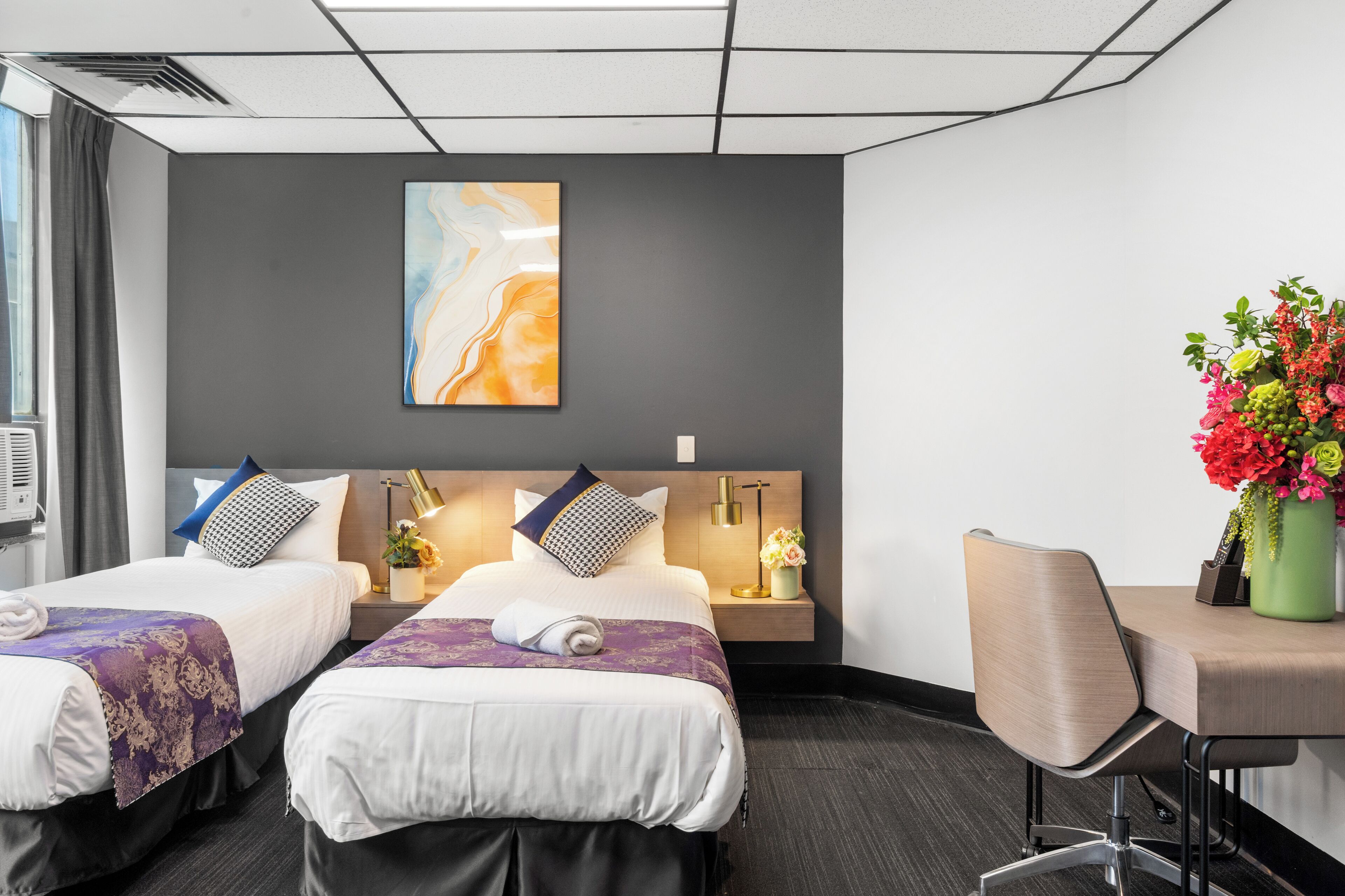 Habitación de hotel con dos camas, un escritorio, una silla, una mesita de noche con lámpara, un florero con flores y un cuadro en la pared.