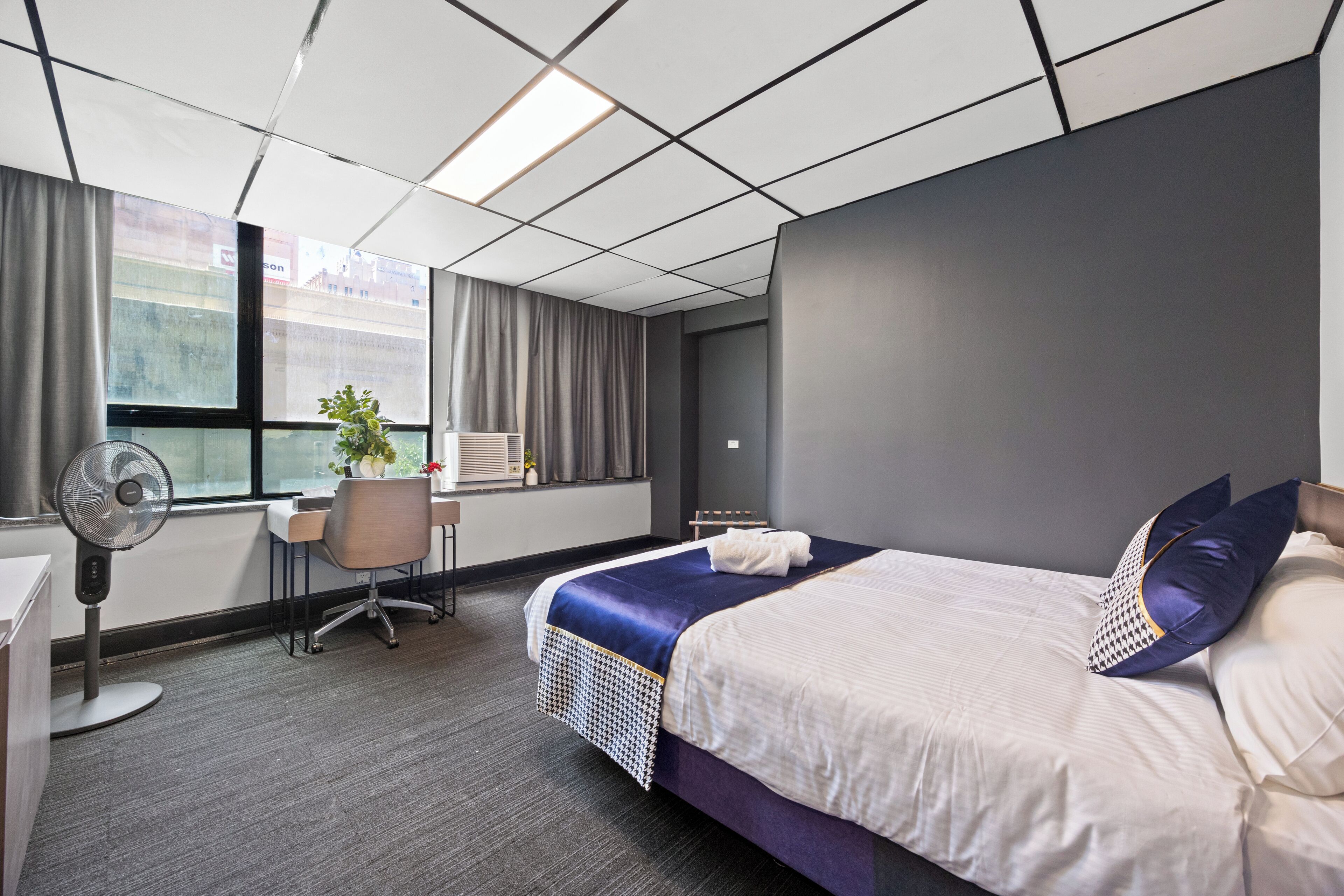 A hotel room with a bed, a desk, a chair, a fan, and a window with curtains.