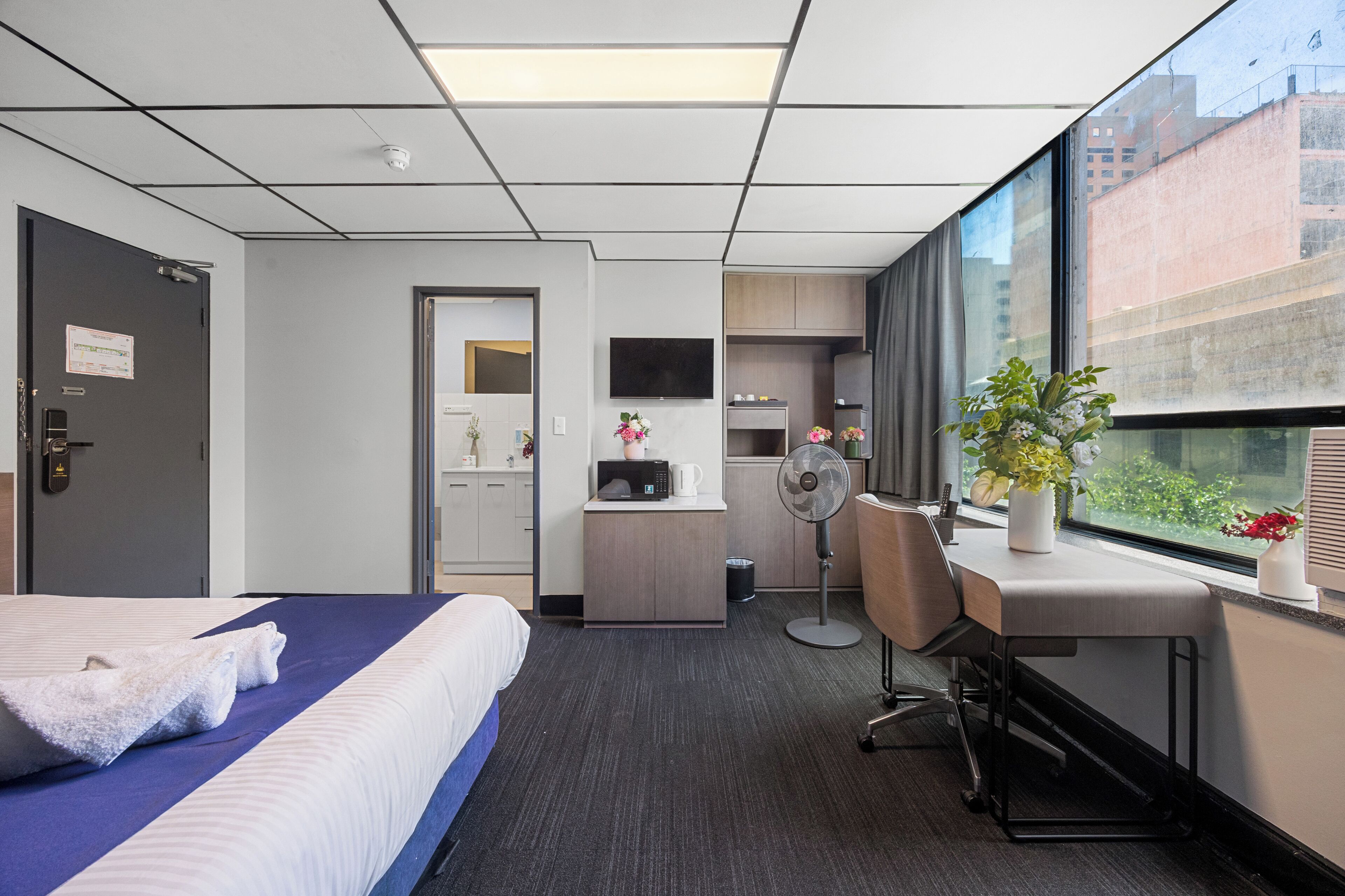Une chambre d’hôtel équipée d’un lit, d’un bureau, d’une chaise, d’un ventilateur, d’un four à micro-ondes, d’une petite kitchenette et d’une grande fenêtre donnant sur des immeubles.