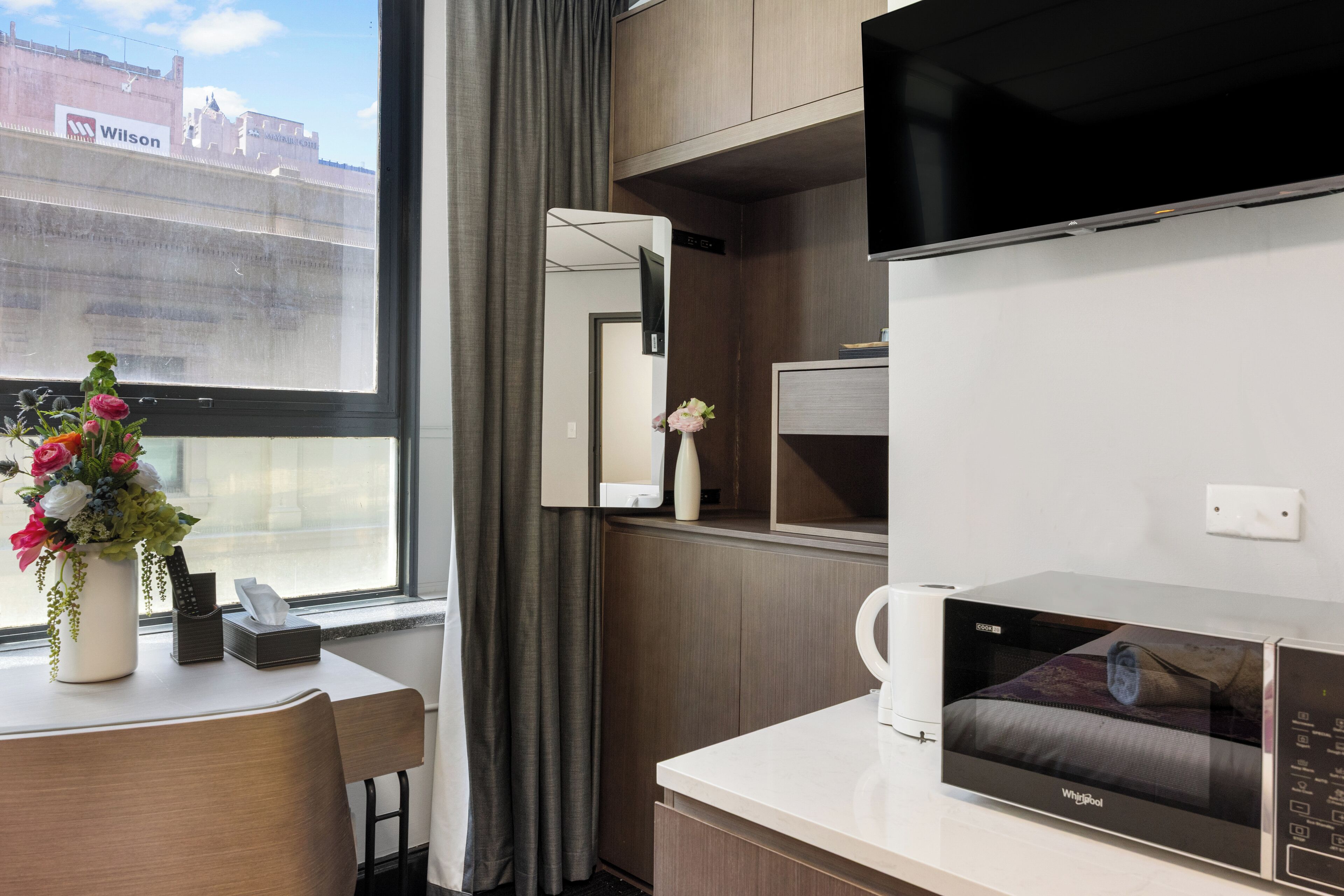 Ein Hotelzimmer mit Schreibtisch, Mikrowelle, Fernseher, einem Fenster mit Blick auf ein Gebäude und einer Vase mit Blumen.