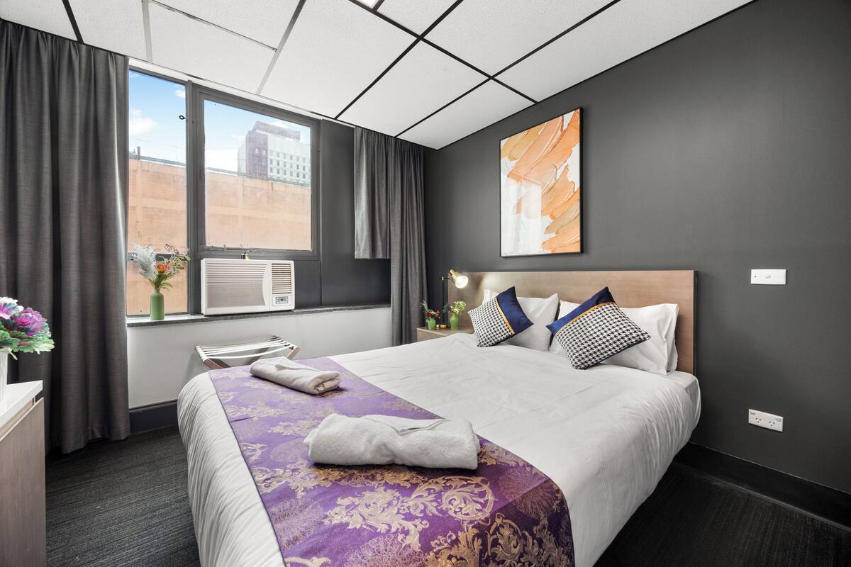 A hotel room with a large bed, a window with curtains, a wall-mounted artwork, and a bedside table with a lamp.