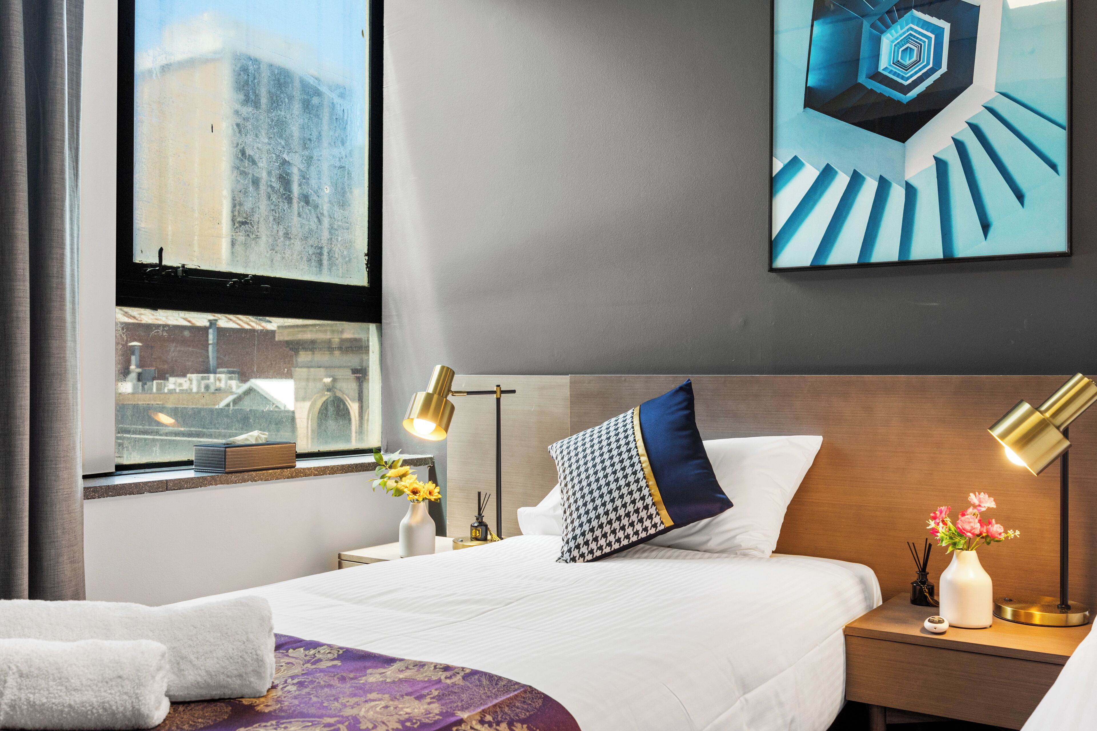 A hotel room with a large bed, a nightstand with a lamp, a vase with flowers, and a window with a view of buildings.