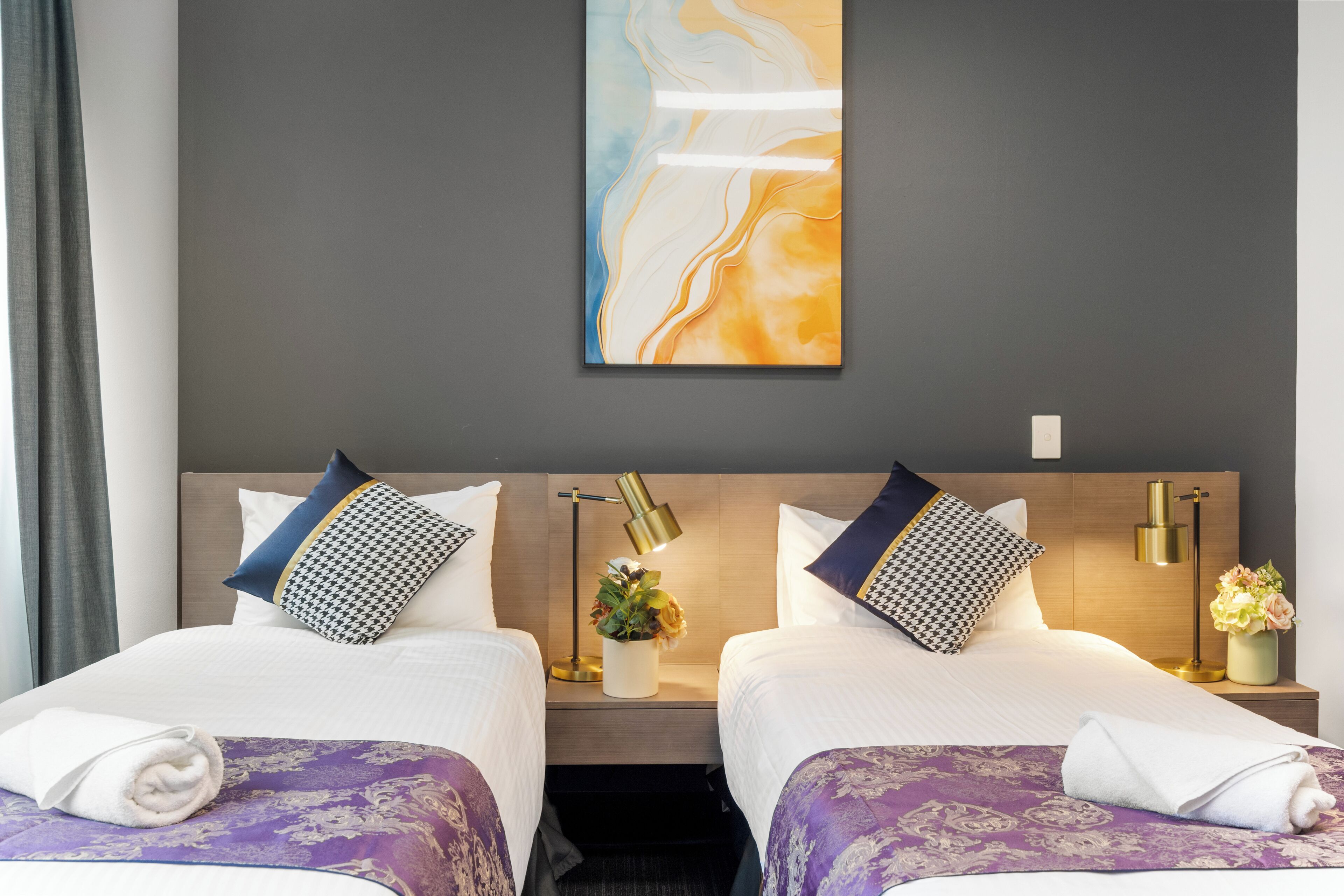 A hotel room with two beds, a marble wall art piece, a nightstand with a lamp, and a vase with flowers.