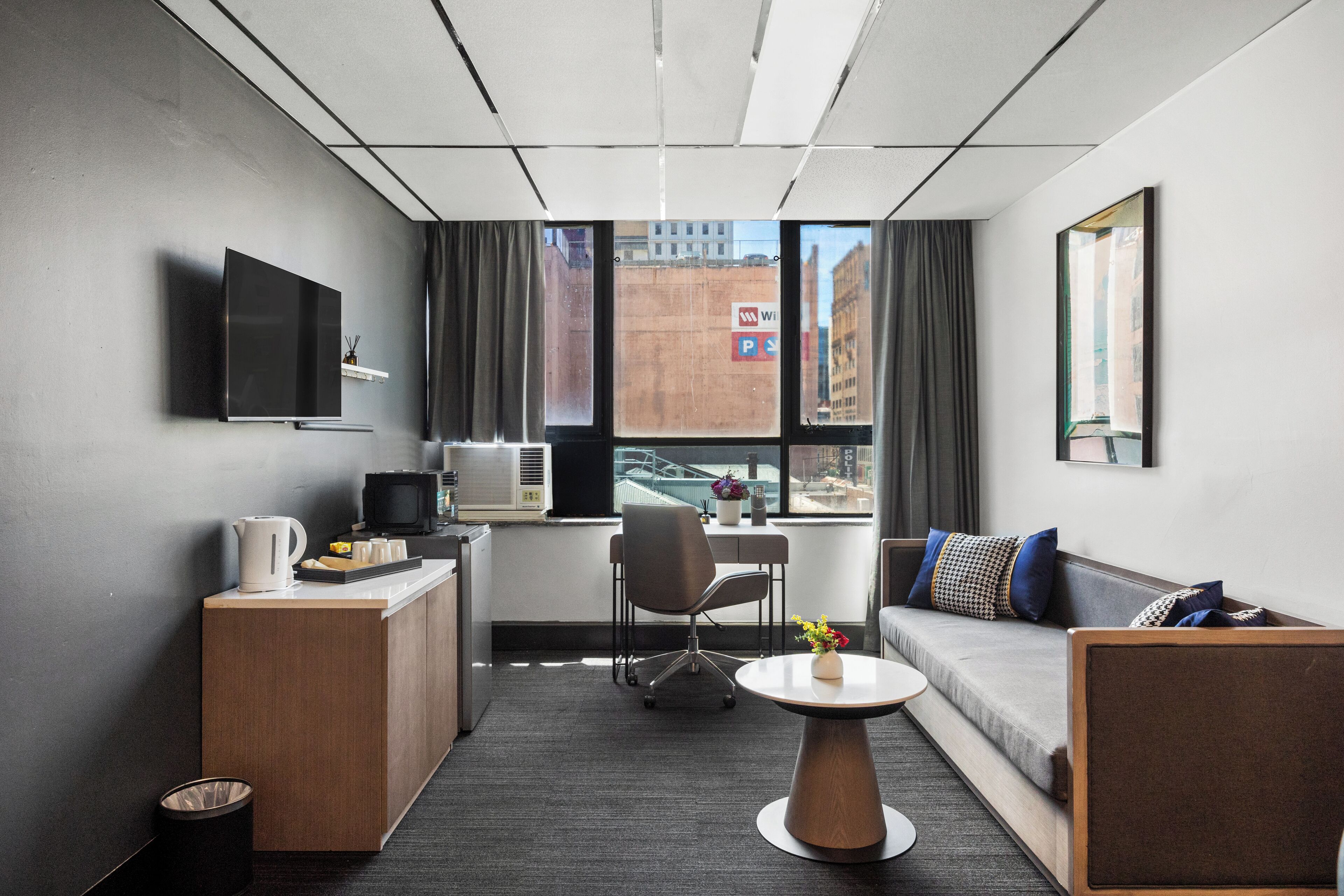 A modern hotel room with a sofa, a desk, a TV, and a view of the city.