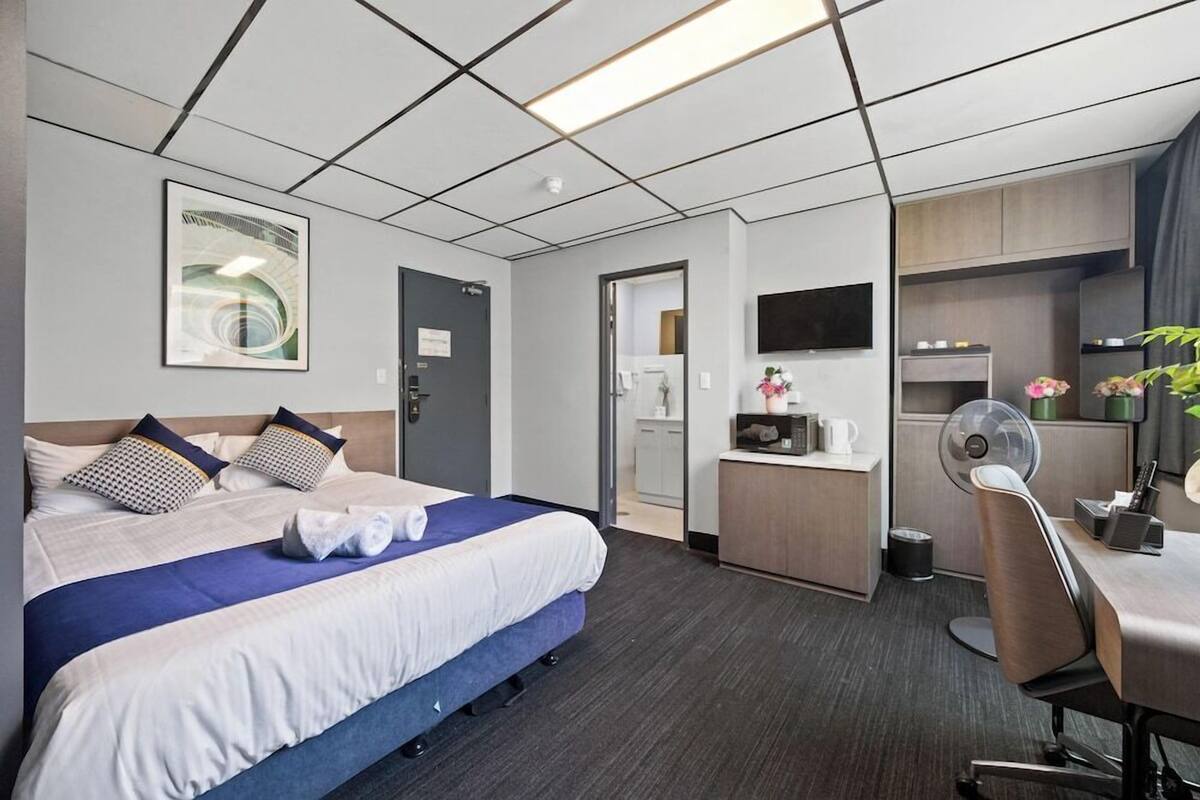A hotel room with a bed, a desk, a chair, a TV, a small kitchenette, and a bathroom.