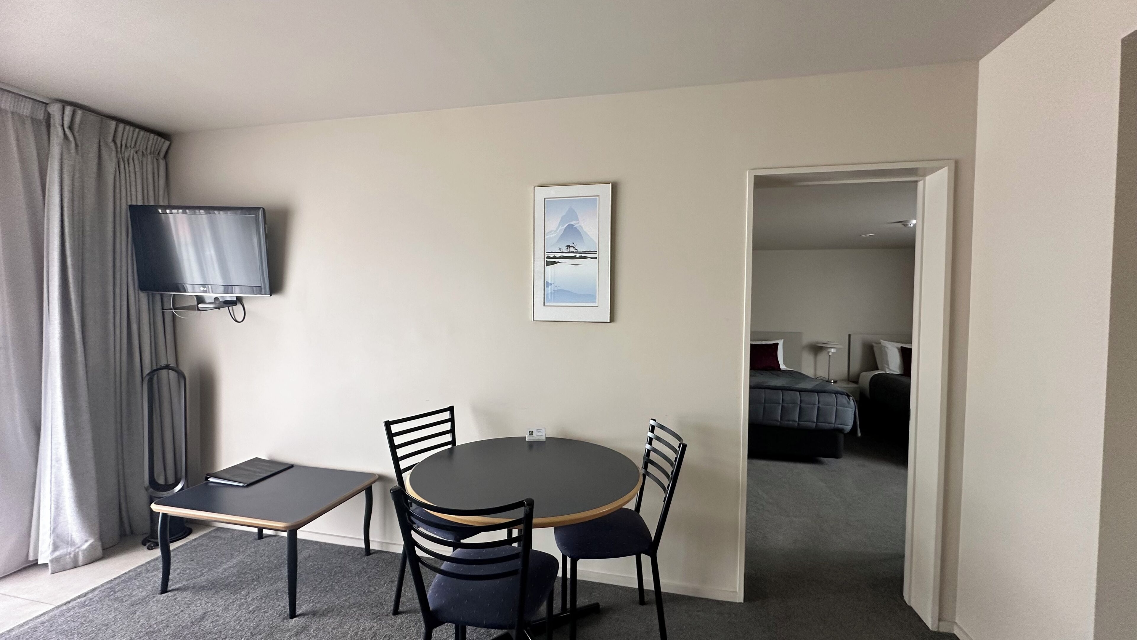 A hotel room with a round table, two chairs, a TV mounted on the wall, and a framed picture above the table.