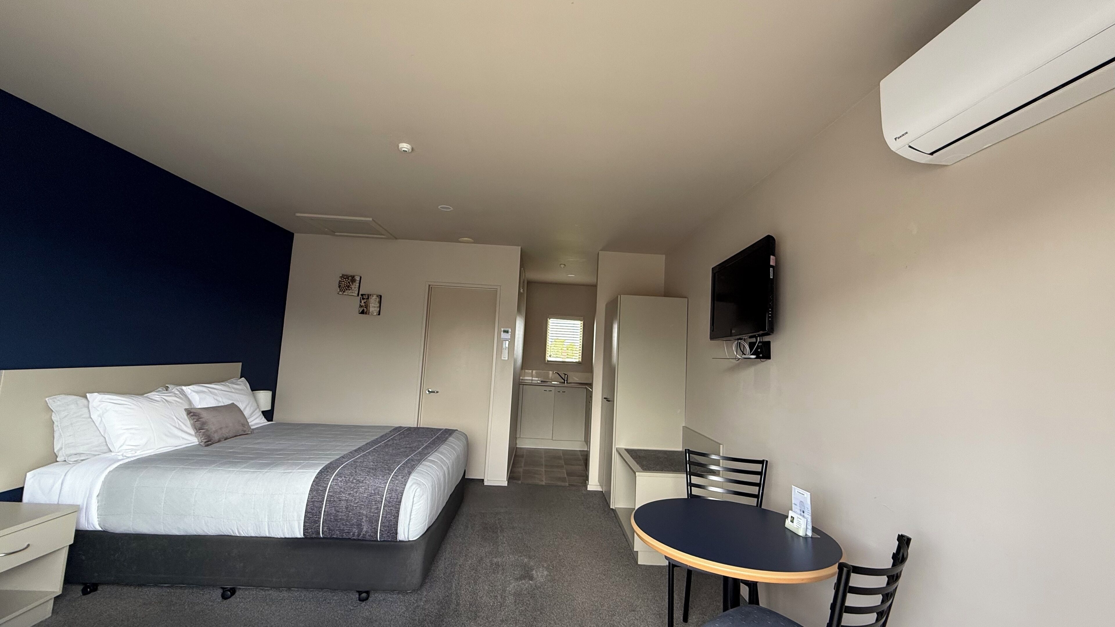 A hotel room with a bed, a desk, a chair, a TV, and a kitchenette.