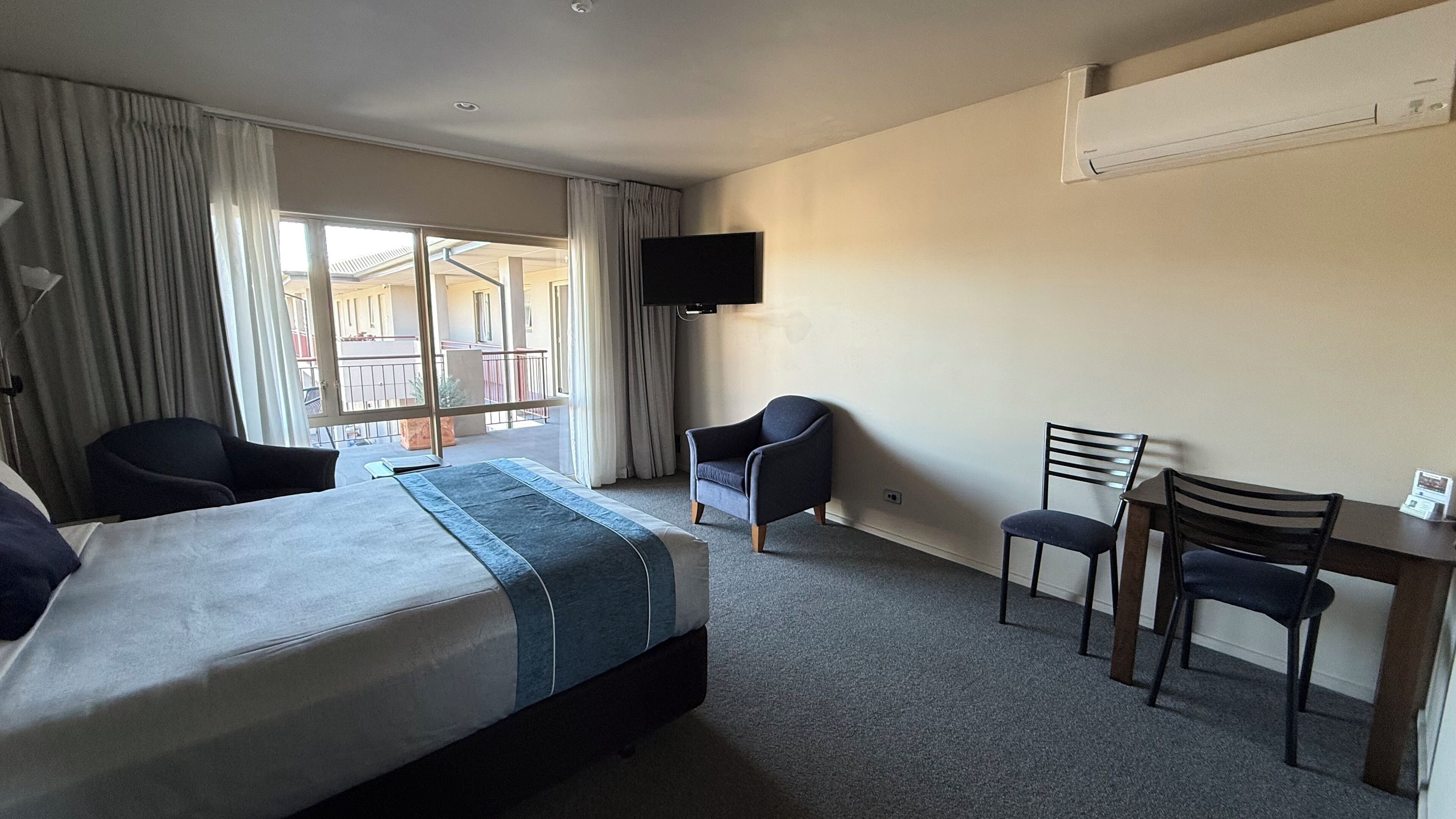 Habitación de hotel con cama, dos sillas, escritorio, televisor y balcón con cortinas.