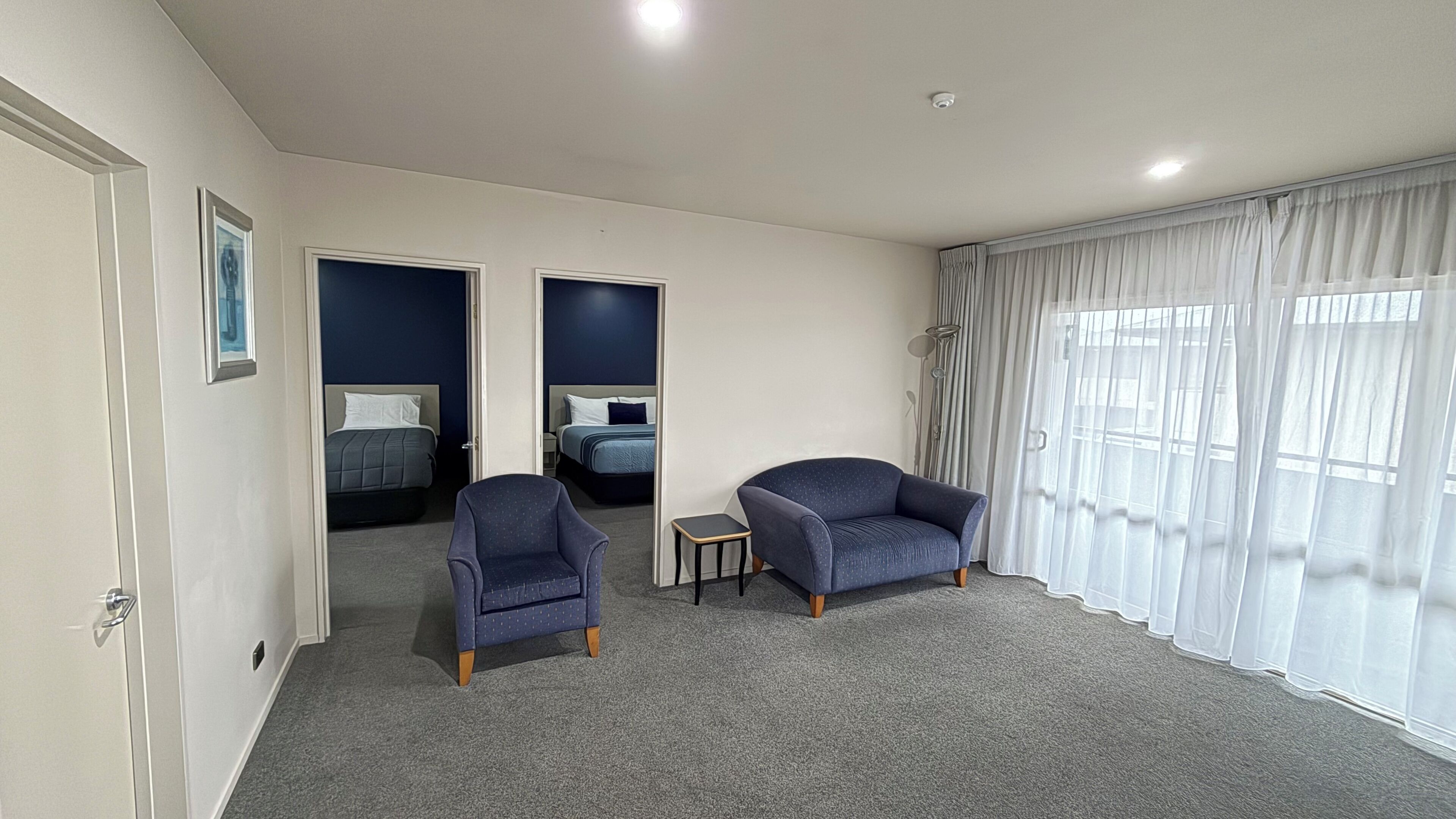 Une chambre d’hôtel avec deux lits, une petite table, deux fauteuils, une fenêtre avec des rideaux légers et une porte ornée d’un cadre de photo.