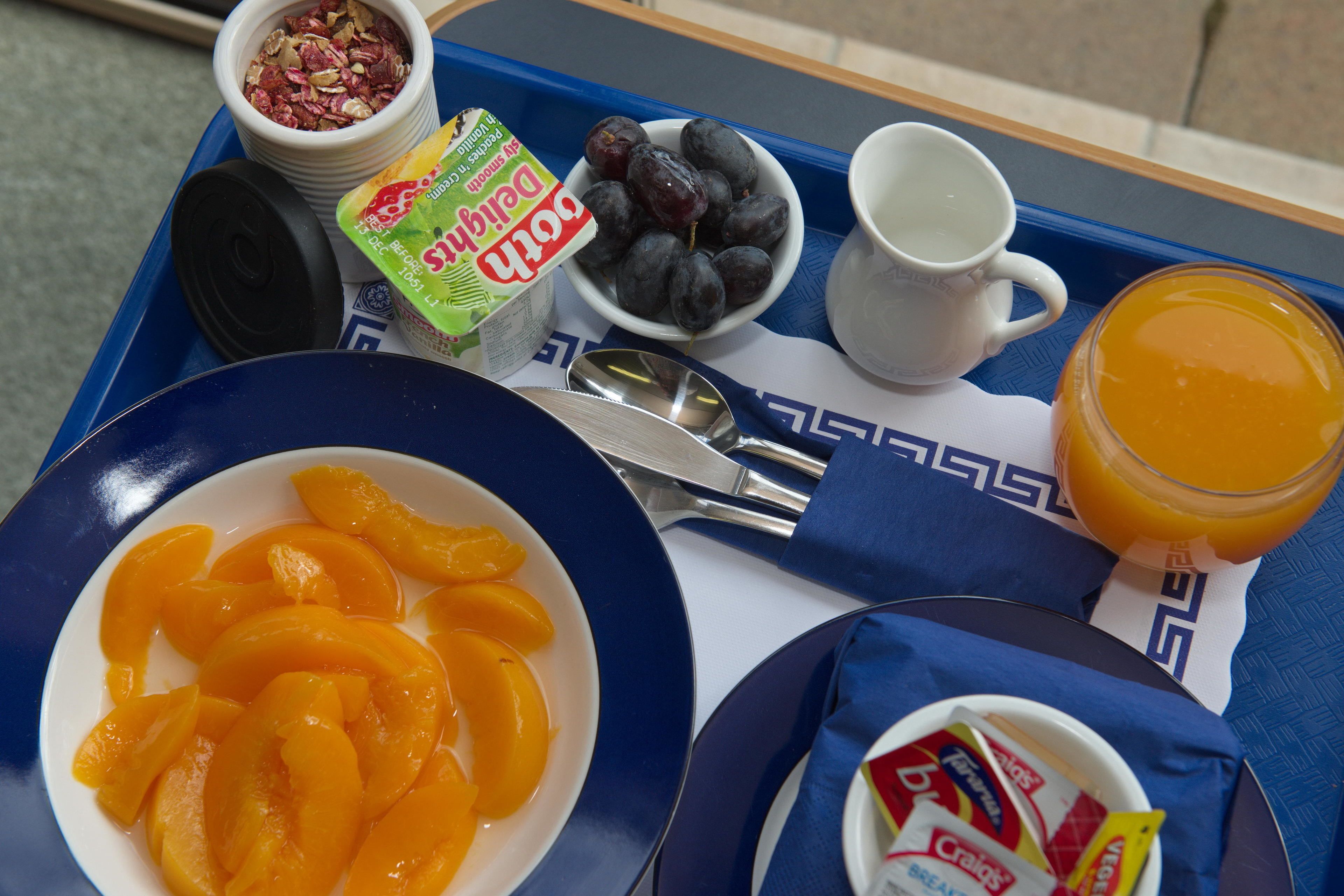 A breakfast tray with a bowl of sliced peaches, a bowl of grapes, a cup of milk, a glass of orange juice, and a container of yogurt.