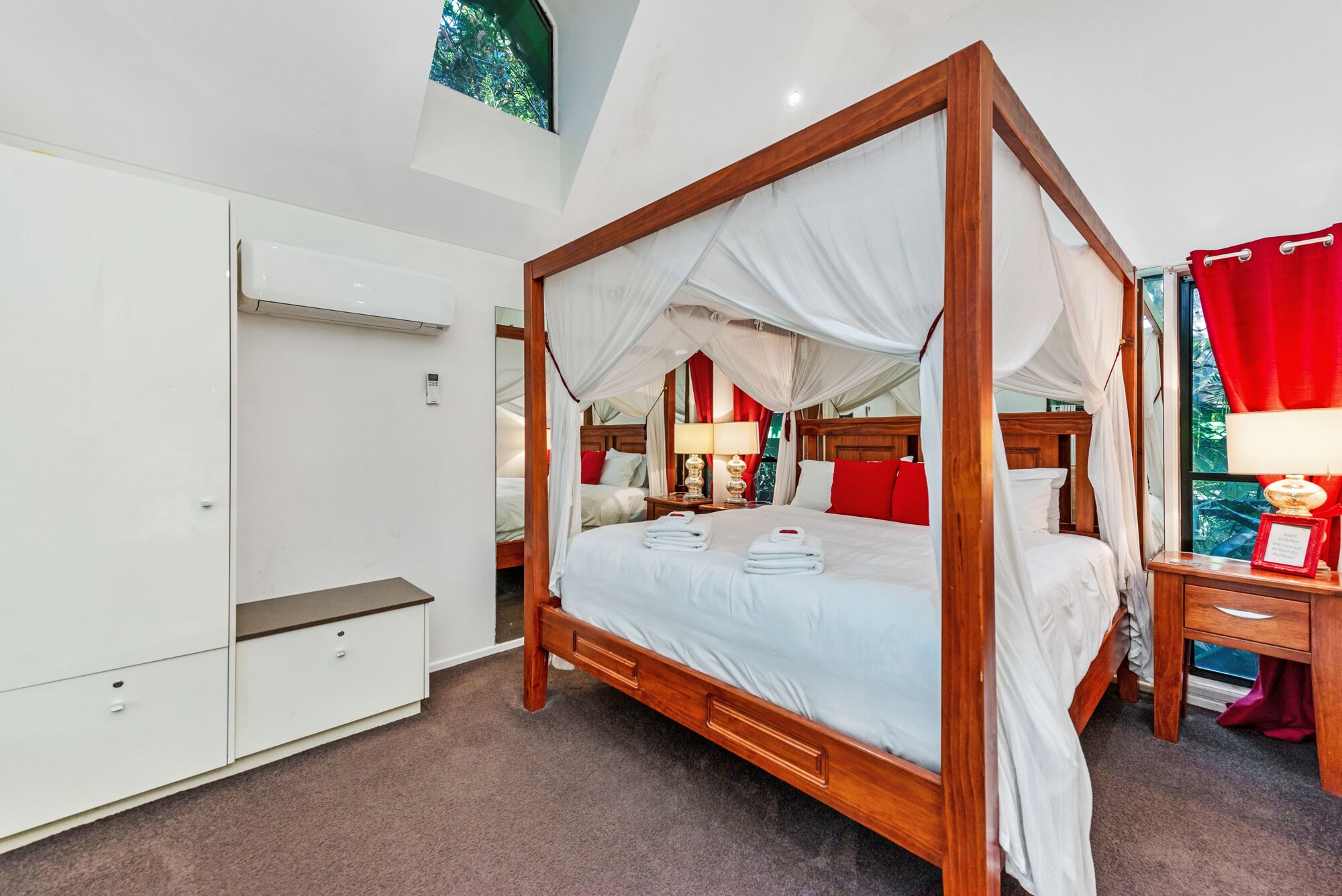 A four-poster canopy bed with white linens, a wooden wardrobe, a bedside table with a lamp, and a red curtain.