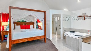 A four-poster bed with white linens and red pillows, a wooden nightstand with a lamp, a bathroom with a bathtub, a large mirror, and a shower. - Linger A While Chalet (Tamborine Mountain)