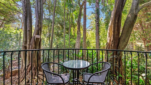 A wooden deck with a glass table and two chairs, surrounded by a metal railing, set in a forested area. - Linger A While Chalet (Tamborine Mountain)