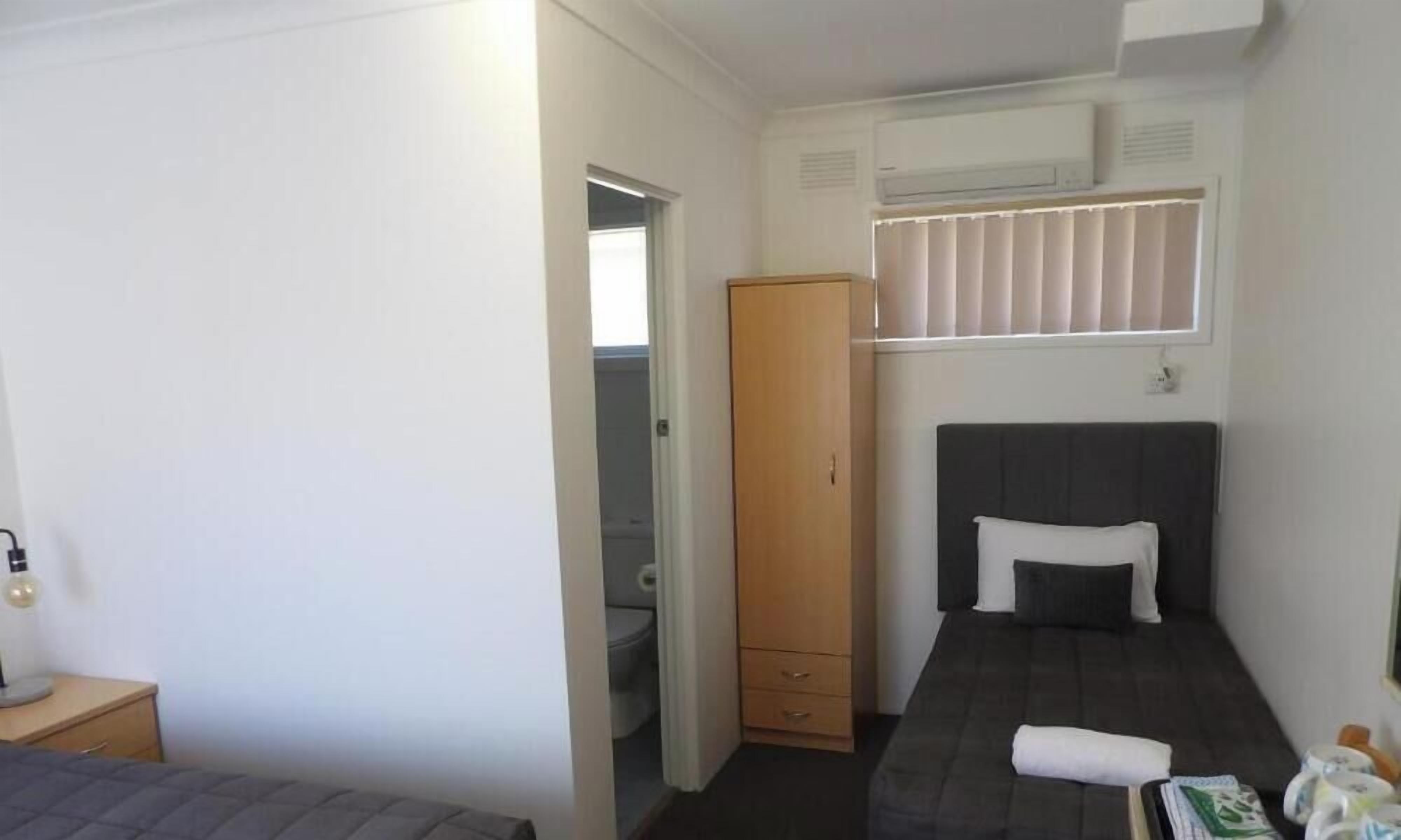 Una habitación pequeña y funcional con una cama individual, un armario, una ventana con persianas y un inodoro.