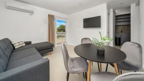 A modern living room with a sofa, dining table, chairs, a potted plant, a flat-screen TV, and a sliding door leading to a balcony. - Kiama Motel 617 (Kiama)