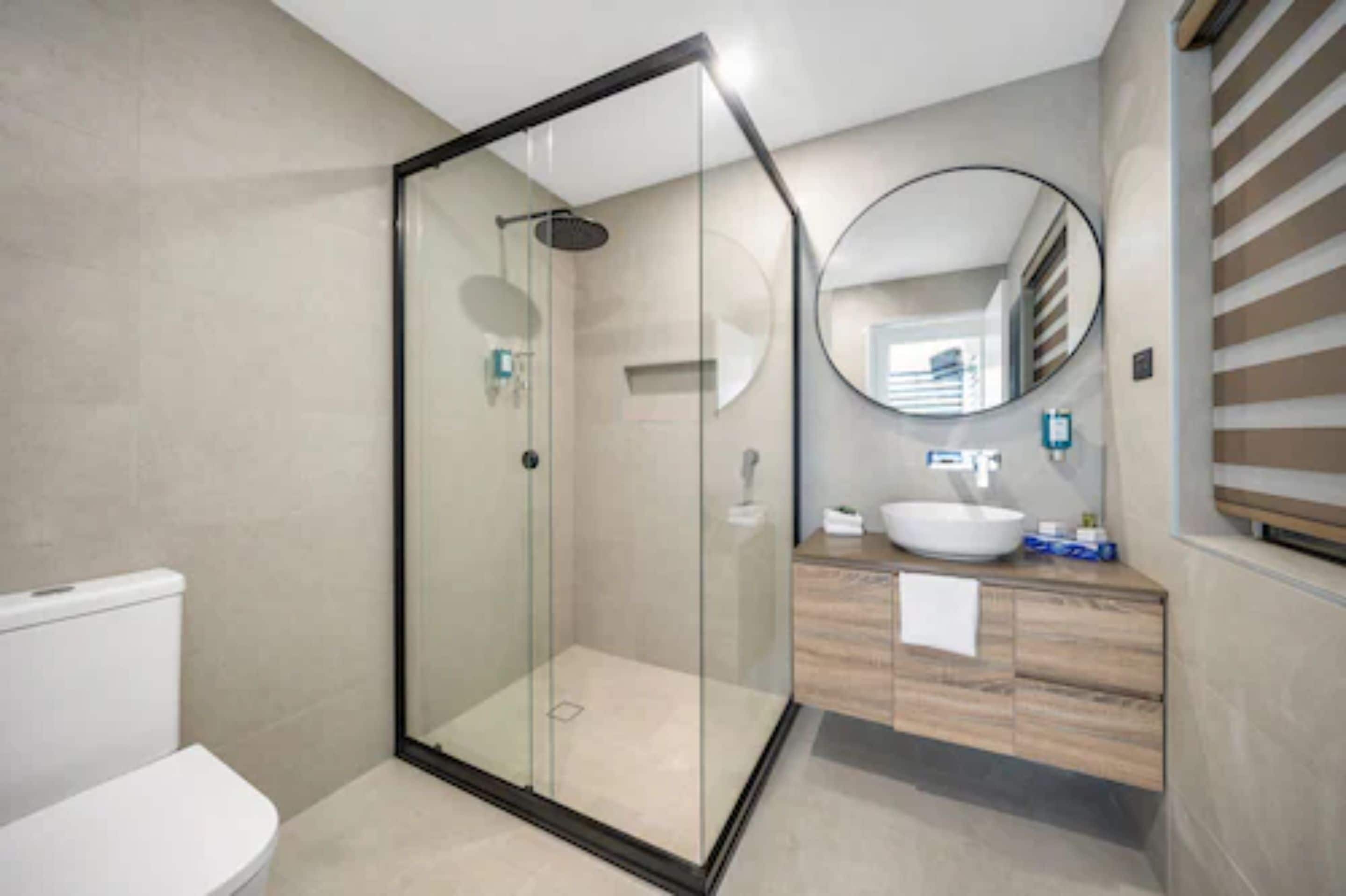 Une salle de bain moderne avec une douche à parois de verre, des toilettes blanches, un meuble-lavabo en bois et un miroir rond.