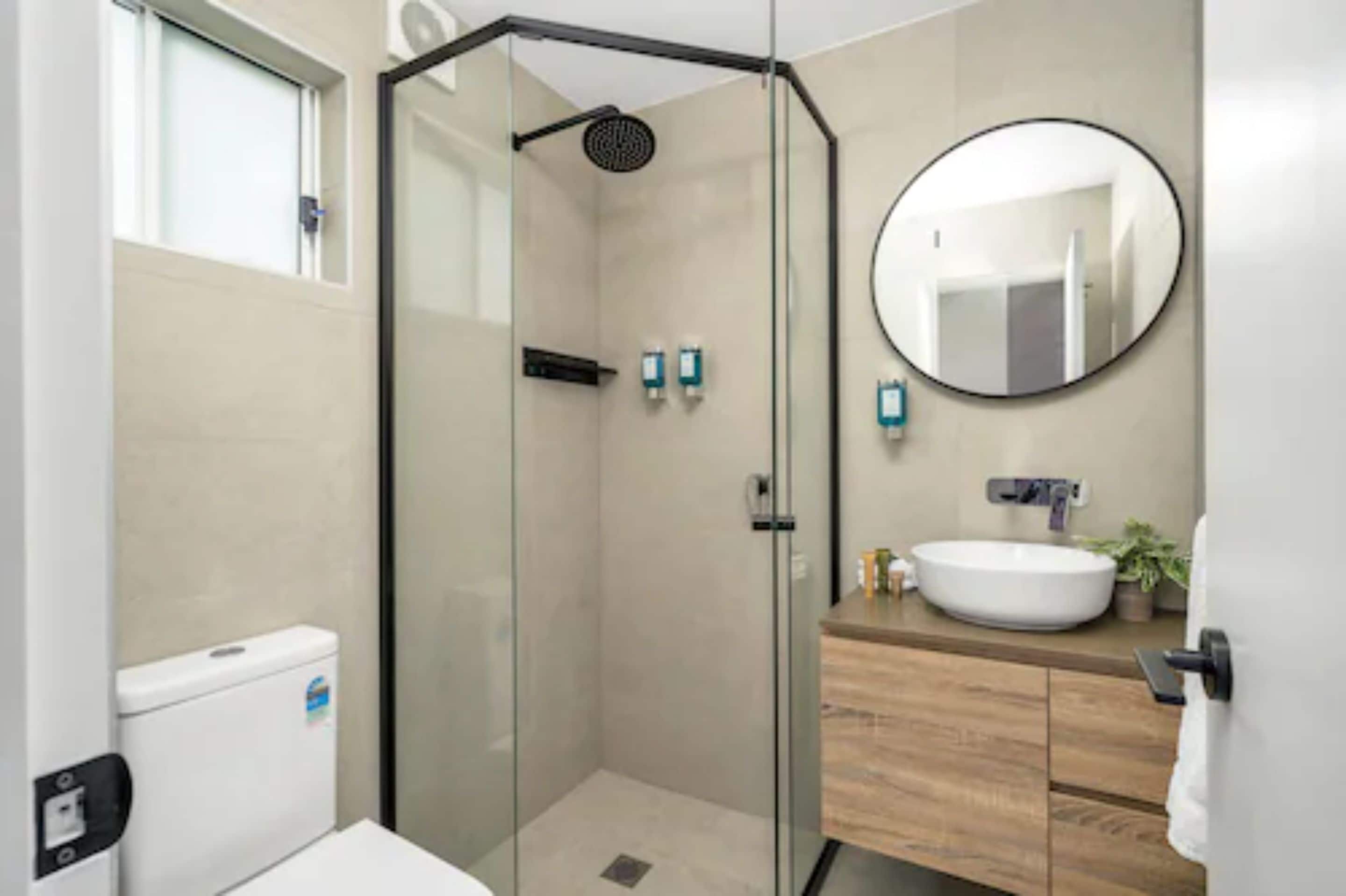 A modern bathroom with a glass-enclosed shower, a round mirror, and a wooden vanity.