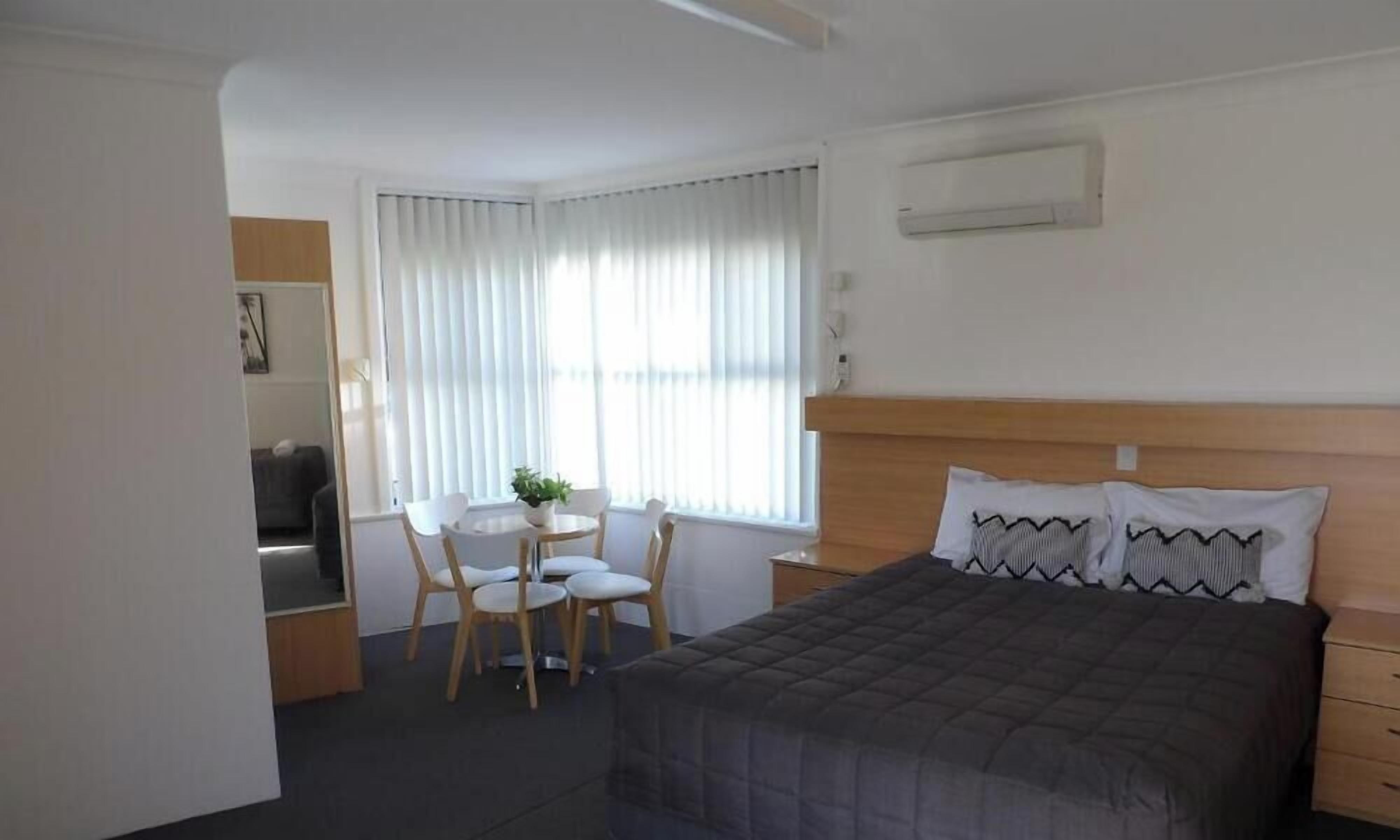 A hotel room with a bed, a desk with a chair, a small table with a plant, and a window with curtains.