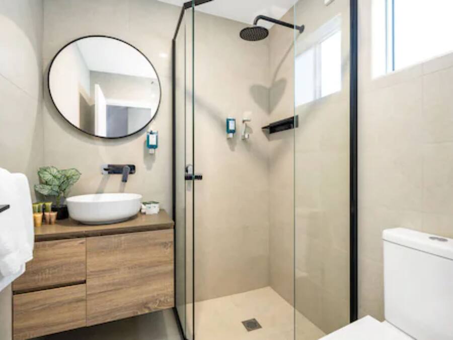 Un baño moderno con ducha envuelta en vidrio, un espejo redondo, un lavamanos blanco y un inodoro.