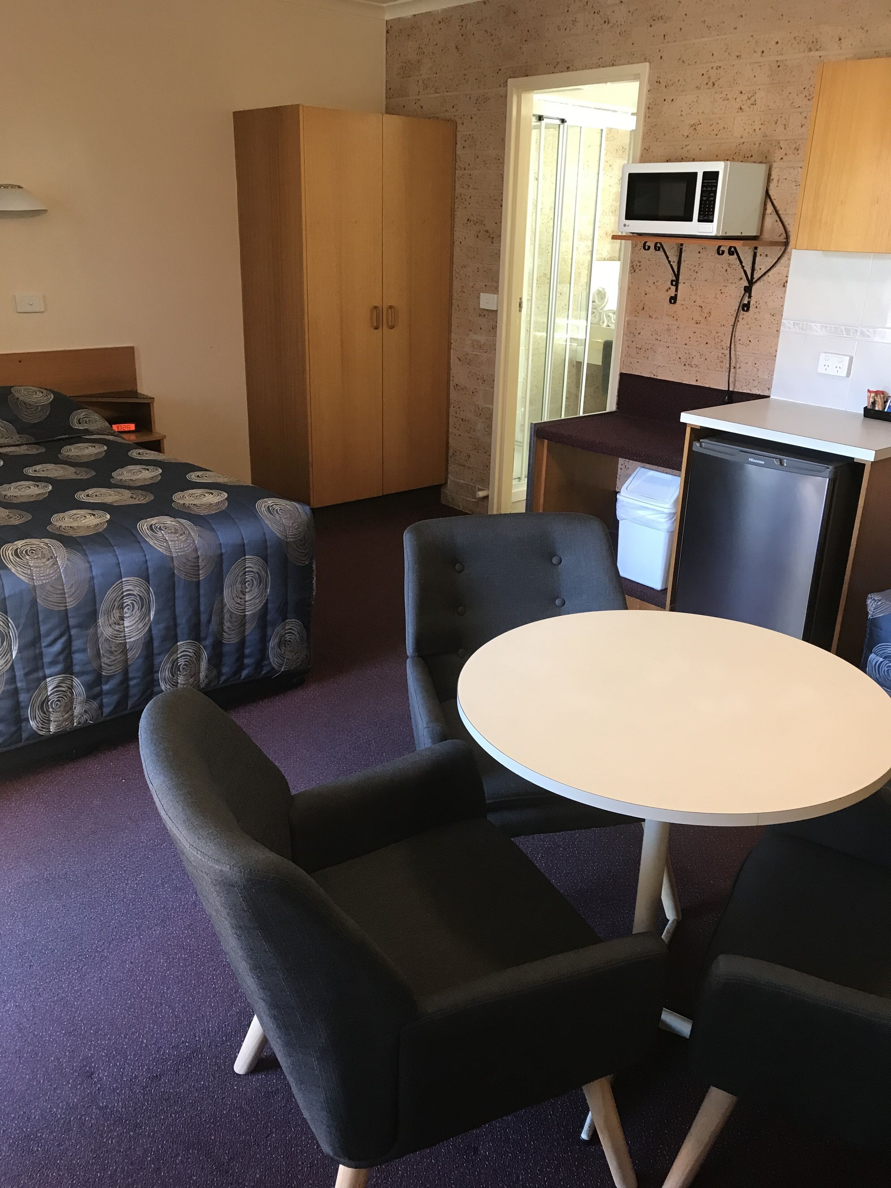 A compact room with a bed, a wardrobe, a small kitchenette with a microwave, and a round table with two chairs.