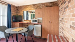 Habitación compacta con pared de ladrillo, televisor, microondas, refrigerador, pequeña mesa de comedor con sillas y un banco a rayas.