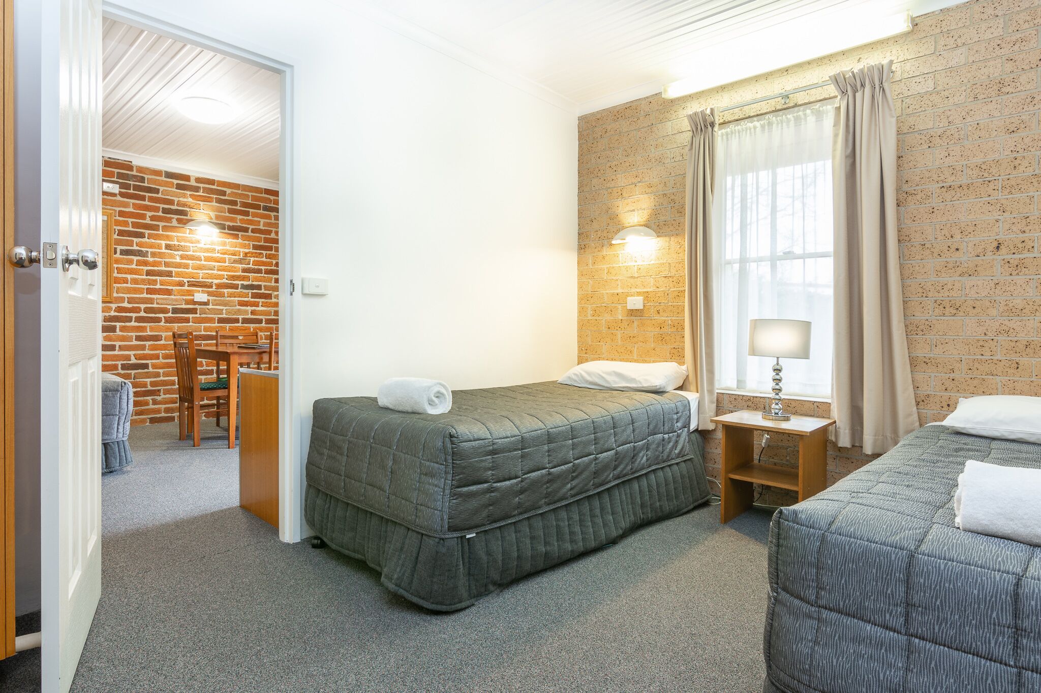 A room with two beds, a brick wall, a window with curtains, and a small table with a lamp.