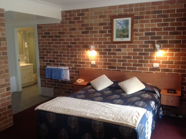 A bedroom with a brick wall, a bed with a patterned duvet, two bedside tables with lamps, a nightstand with a phone, and a framed picture on the wall. - Cedar Lodge Motel (Armidale)