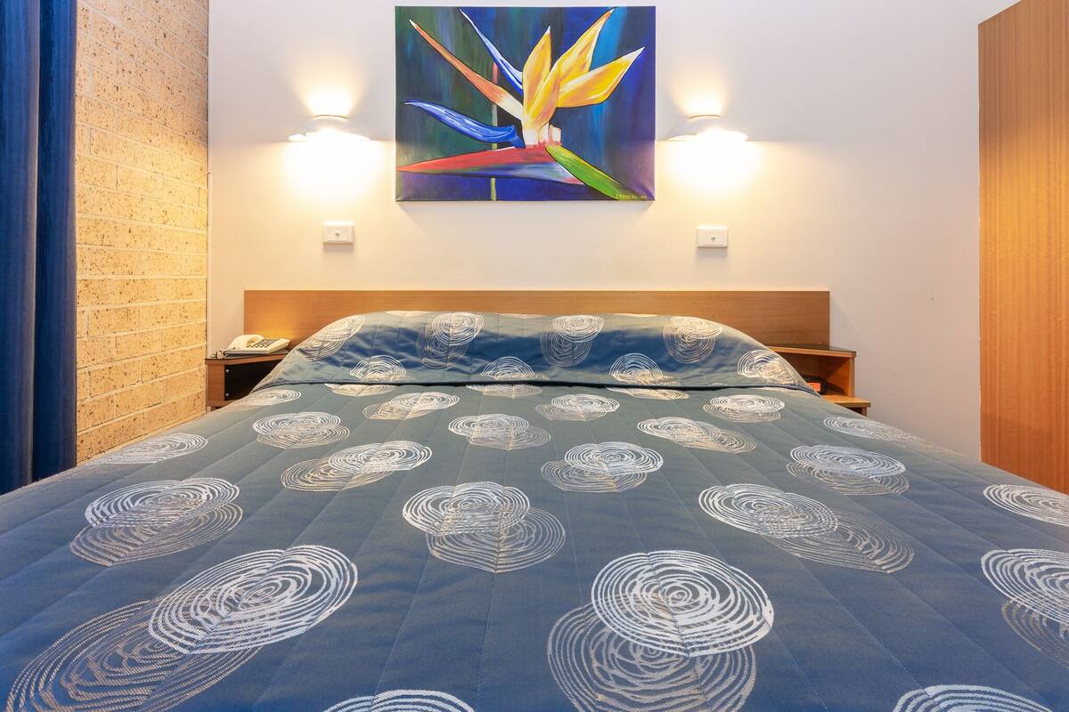 A hotel room with a bed, a painting on the wall, and a brick wall.