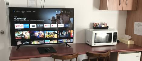 Una kitchenette compacta con microondas, un refrigerador pequeño, un armario de madera y un televisor que muestra la interfaz de un servicio de streaming.