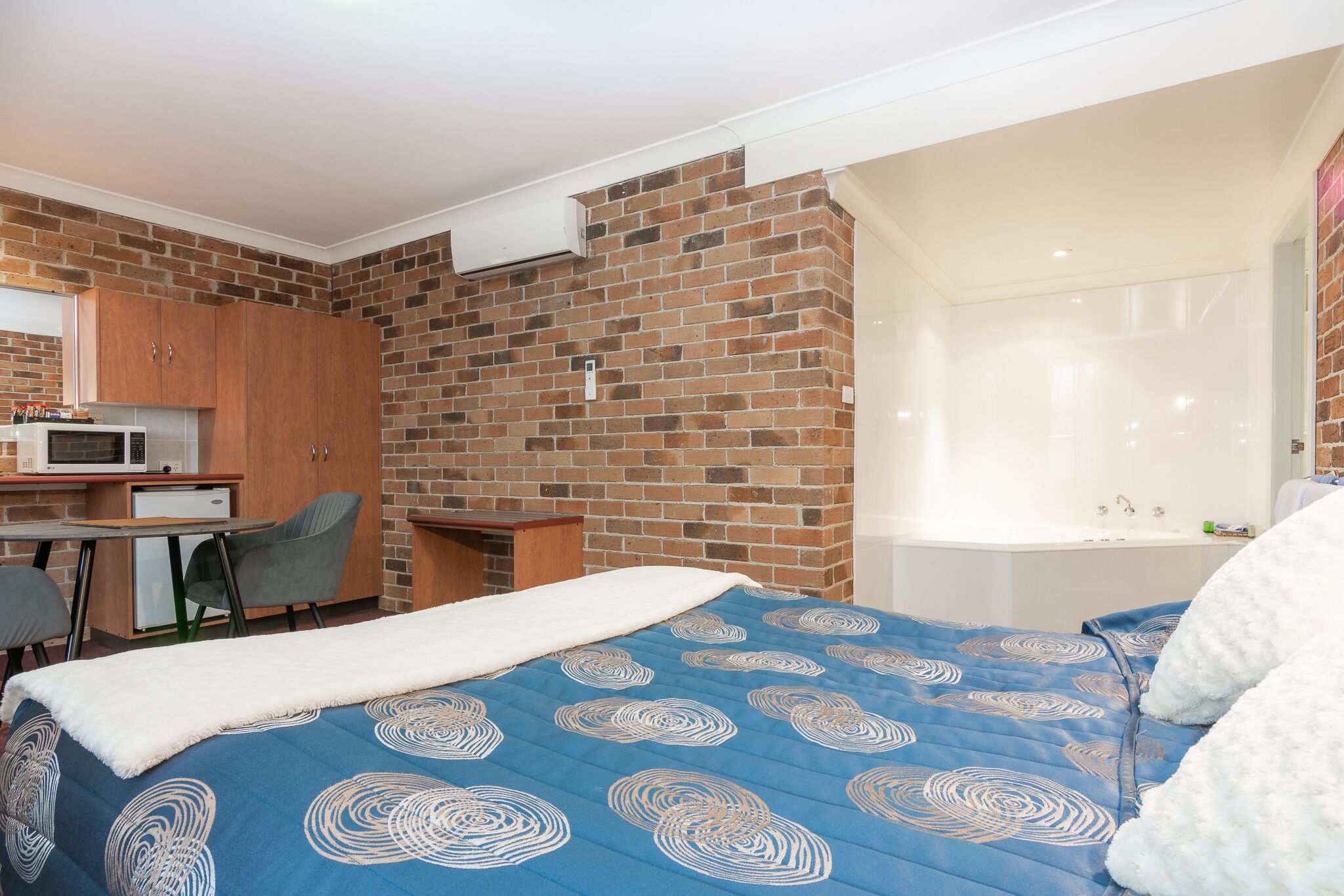 A bedroom with a brick wall, a bed with a blue patterned bedspread, a desk with a microwave, and a small kitchenette.