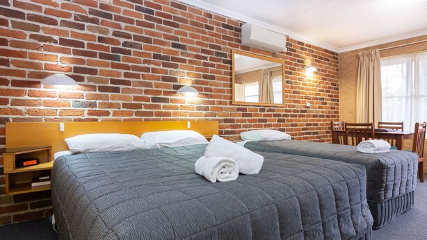 Habitación de hotel con pared de ladrillo, dos camas con colchas grises, una mesita de noche de madera, un escritorio y una silla.