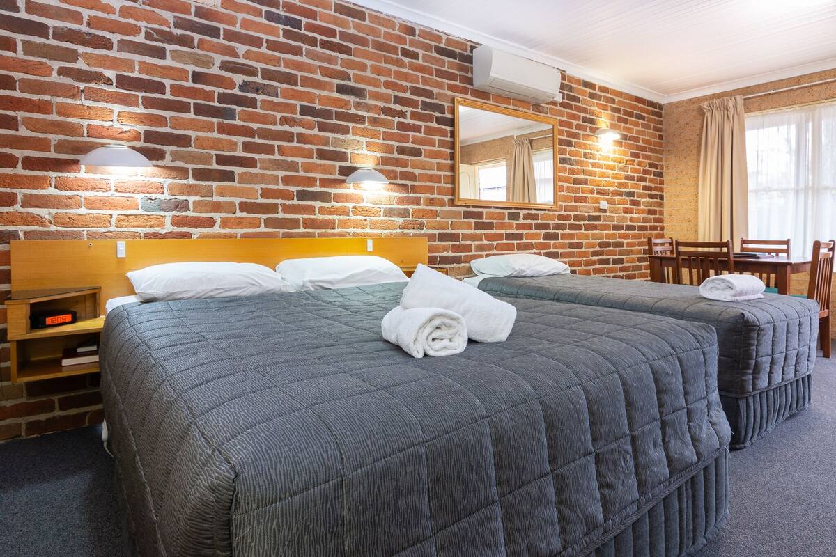 A hotel room with a brick wall, two beds with grey quilts, a wooden nightstand, a desk, and a chair.
