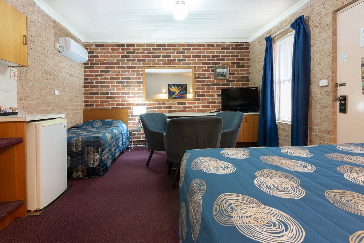 A room with a brick wall, a bed with a blue patterned bedspread, a desk with a chair, a television, and a small refrigerator.